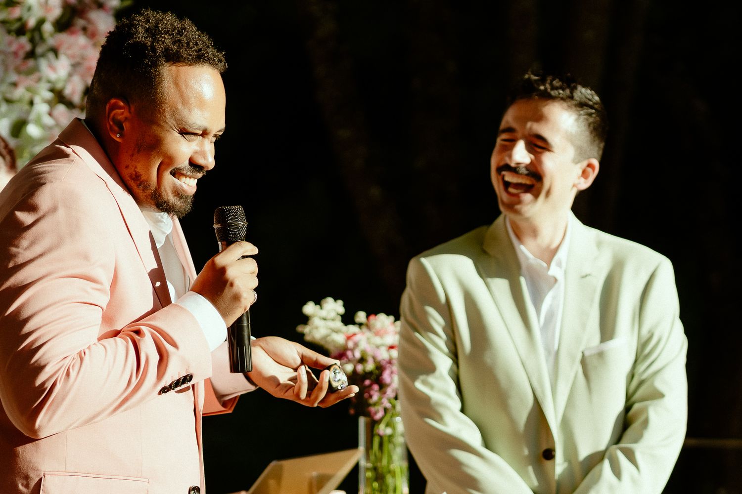 Two men in suits laughing together at a formal event, one holding a microphone, with floral decorations in the background.
