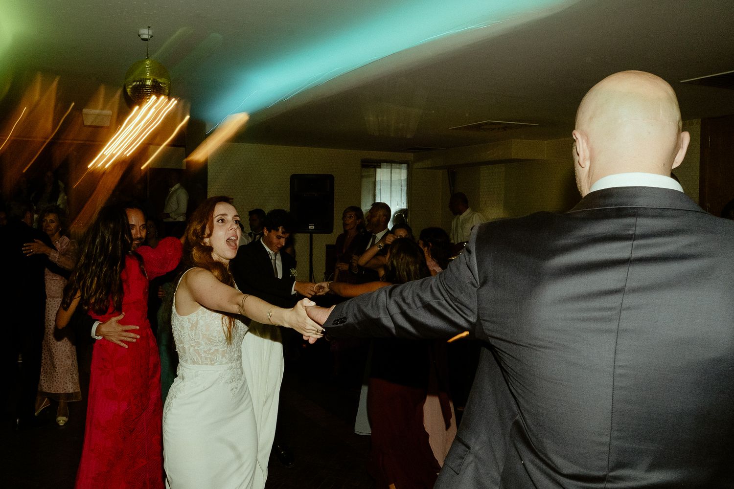 Bride in white dress dancing with guests at a lively wedding reception under colorful lights.