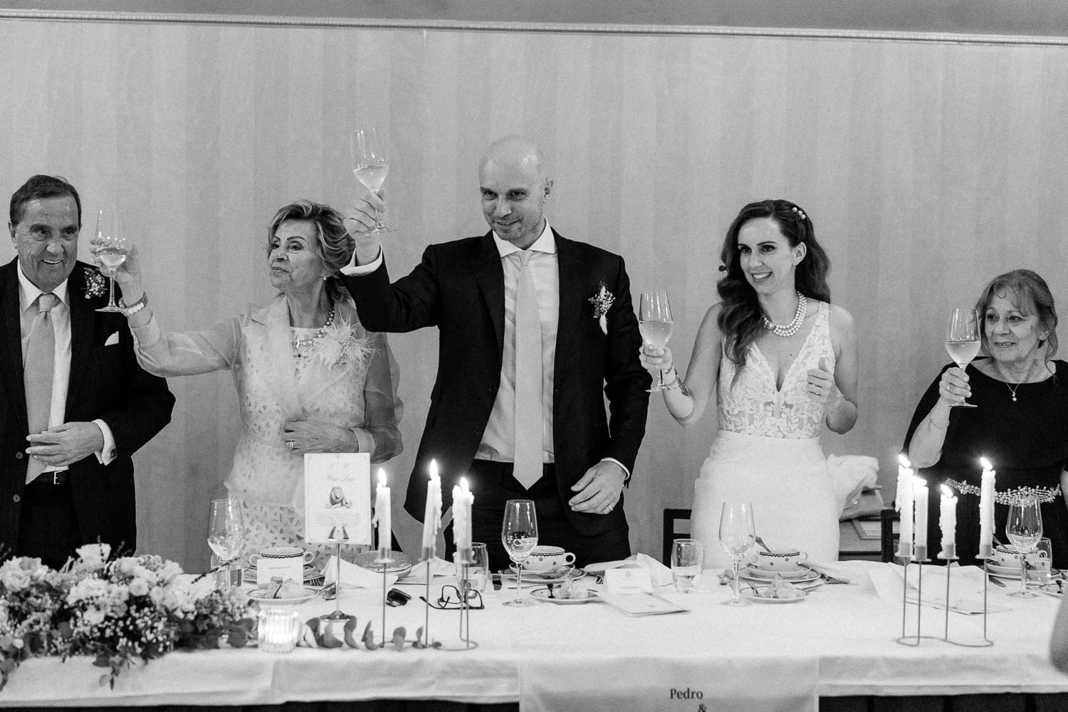 Black and white wedding toast with five people raising champagne glasses at candlelit reception table.