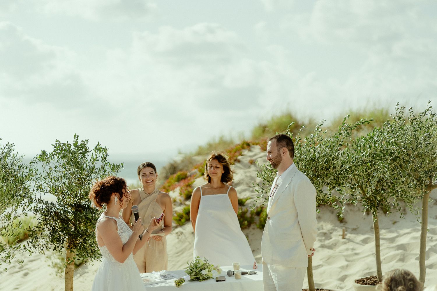 Outdoor beach wedding ceremony on sandy dunes with bride, groom, and guests surrounded by small olive trees.