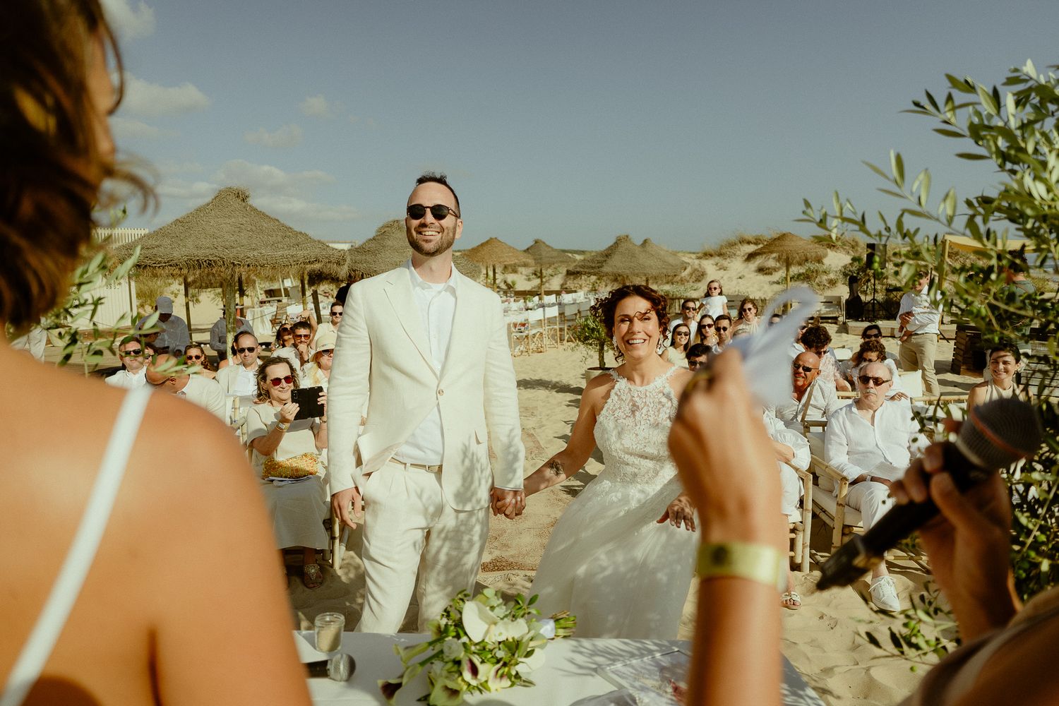 Bride and groom hold hands smiling at outdoor beach wedding ceremony with guests seated on sunny day.