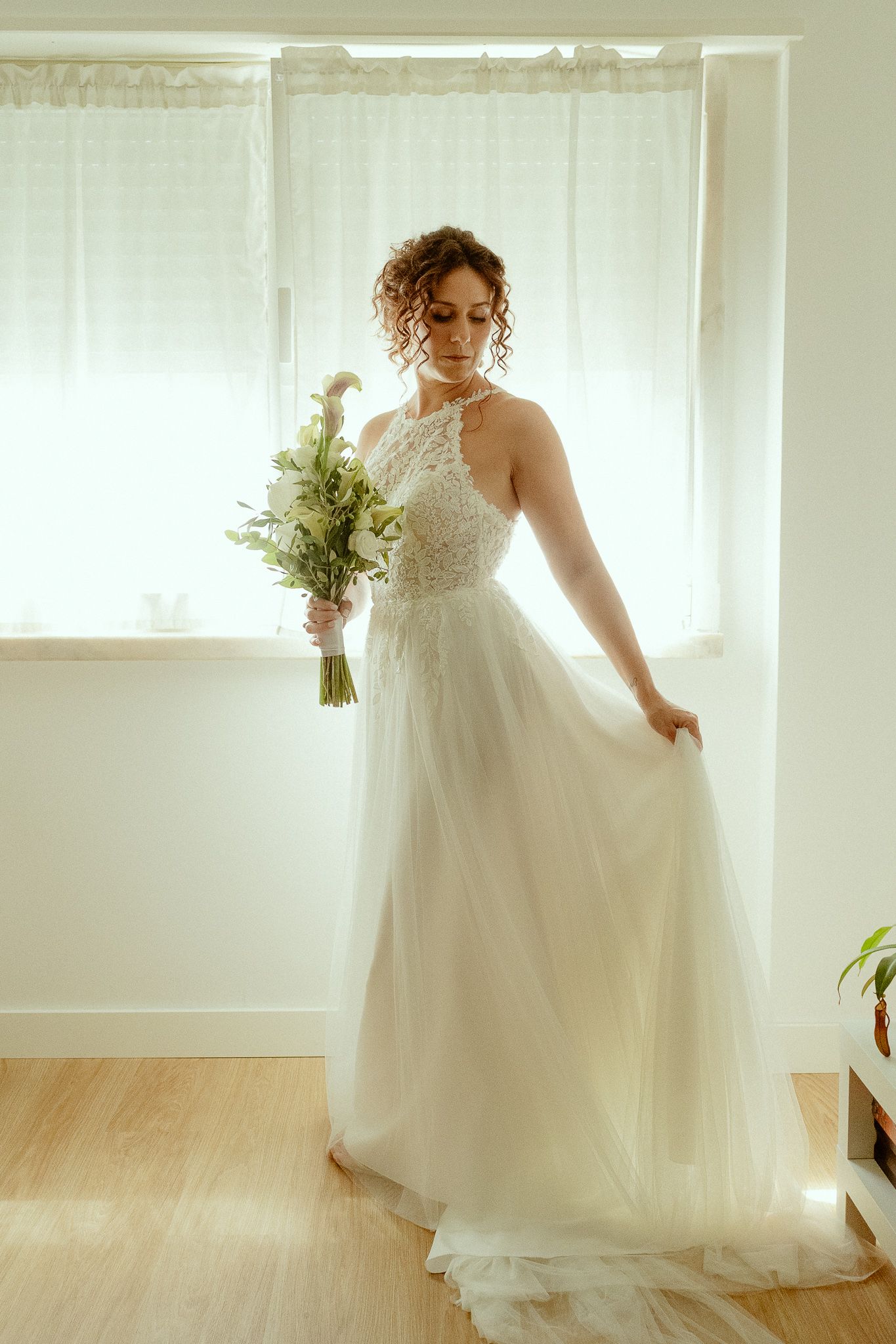 Bride in white lace tulle gown holding green bouquet, standing by window in soft natural light.