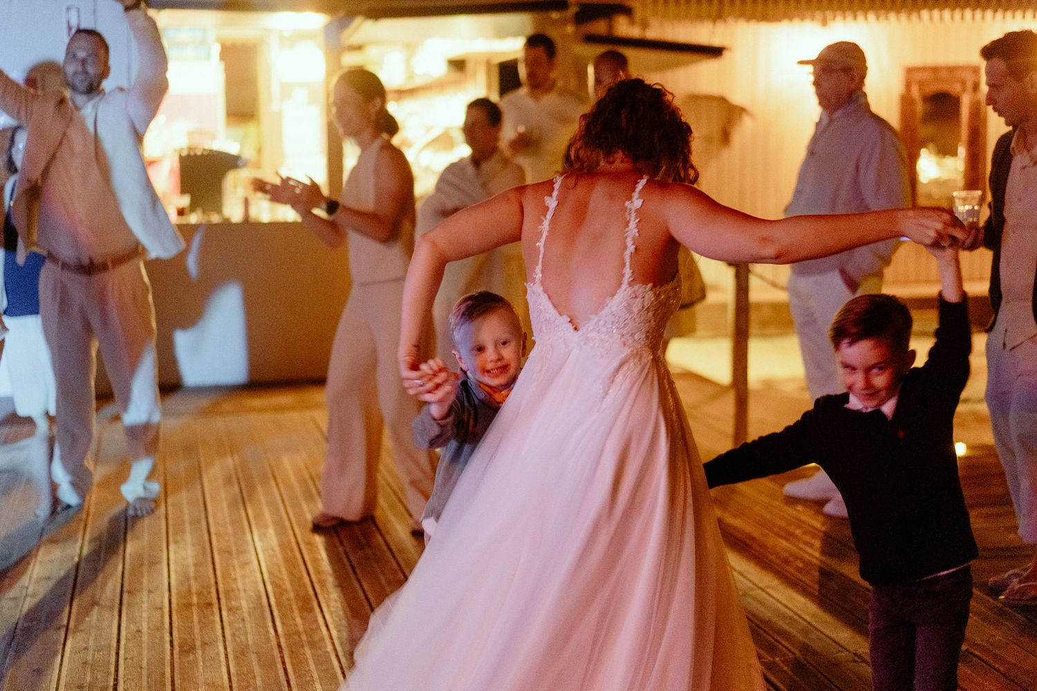 Bride in white gown dancing joyfully with two young boys on wooden dance floor at wedding reception.