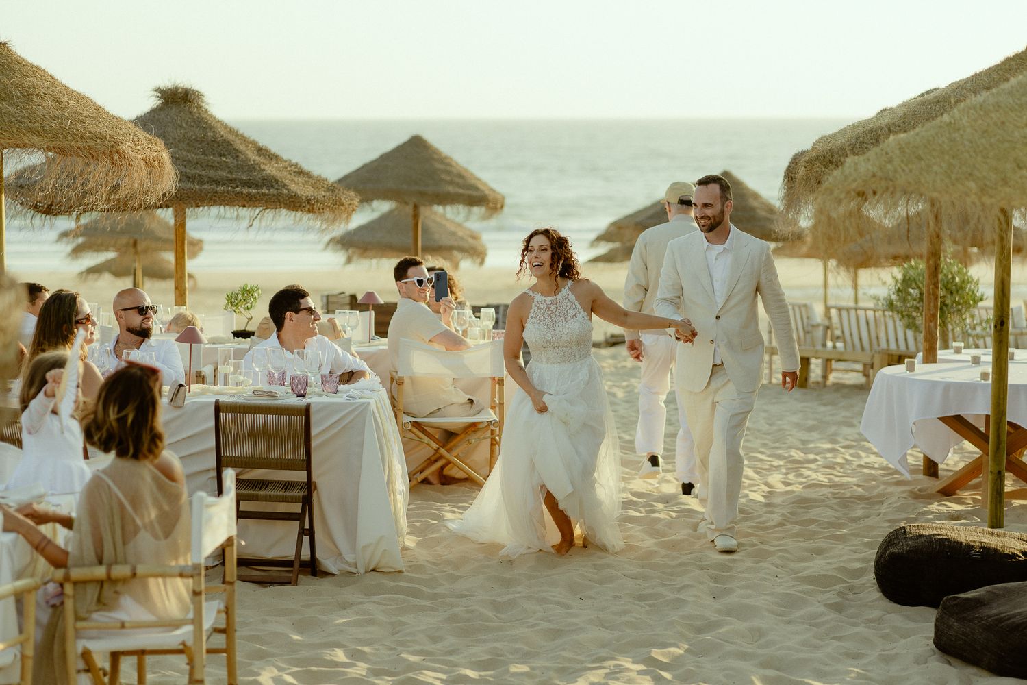 Bride and groom walk joyfully on sandy beach wedding venue with thatched umbrellas and guests in white attire.