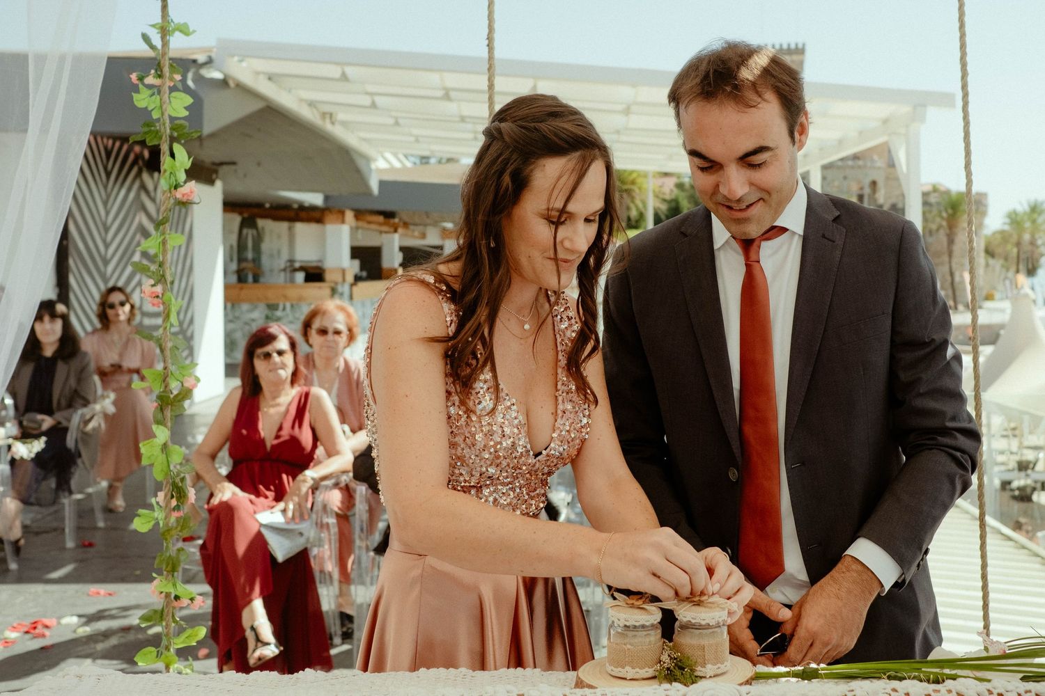 A couple in formal attire serving themselves food at an outdoor event while other guests mingle in the background.