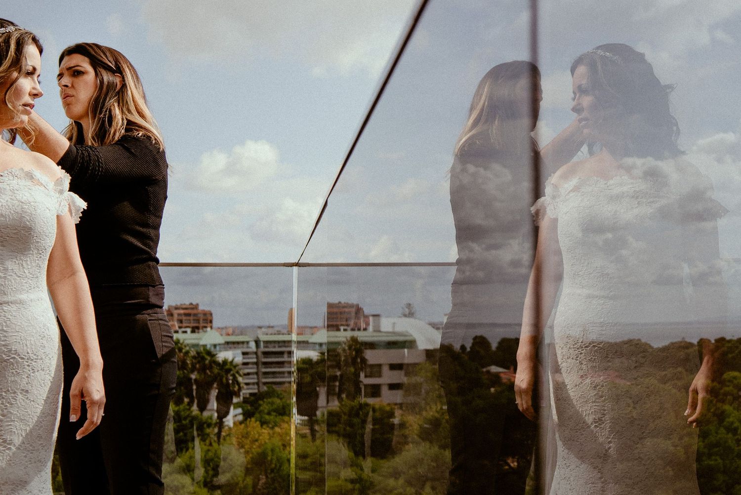 A couple in formal attire poses together on a balcony with a cityscape backdrop during sunset.