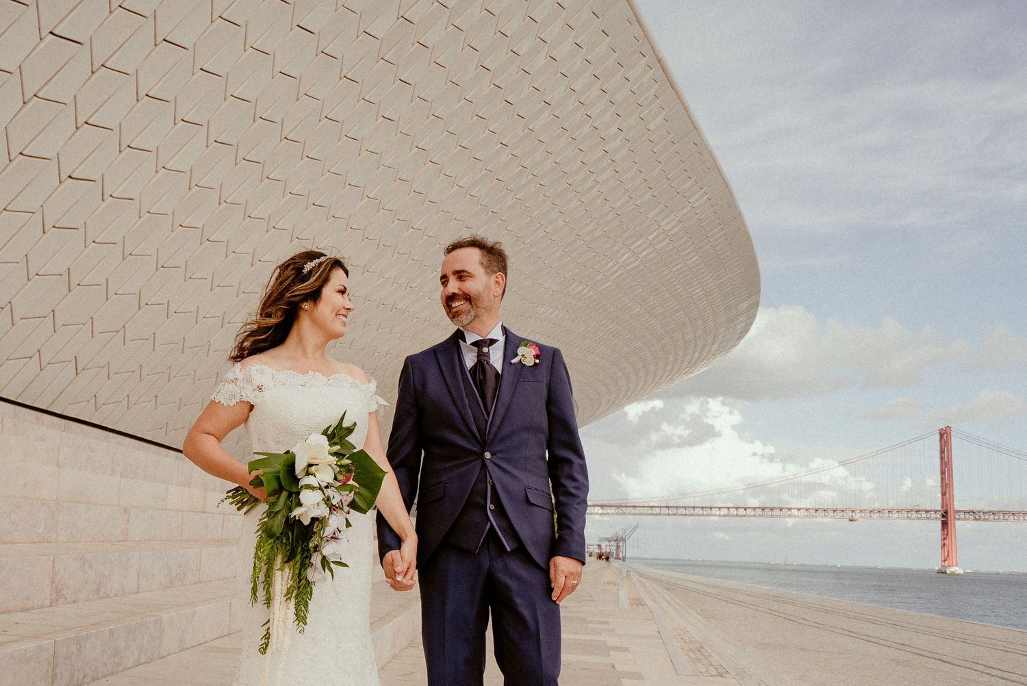 Newlywed couple poses in front of modern white architectural building with Golden Gate Bridge visible in background.
