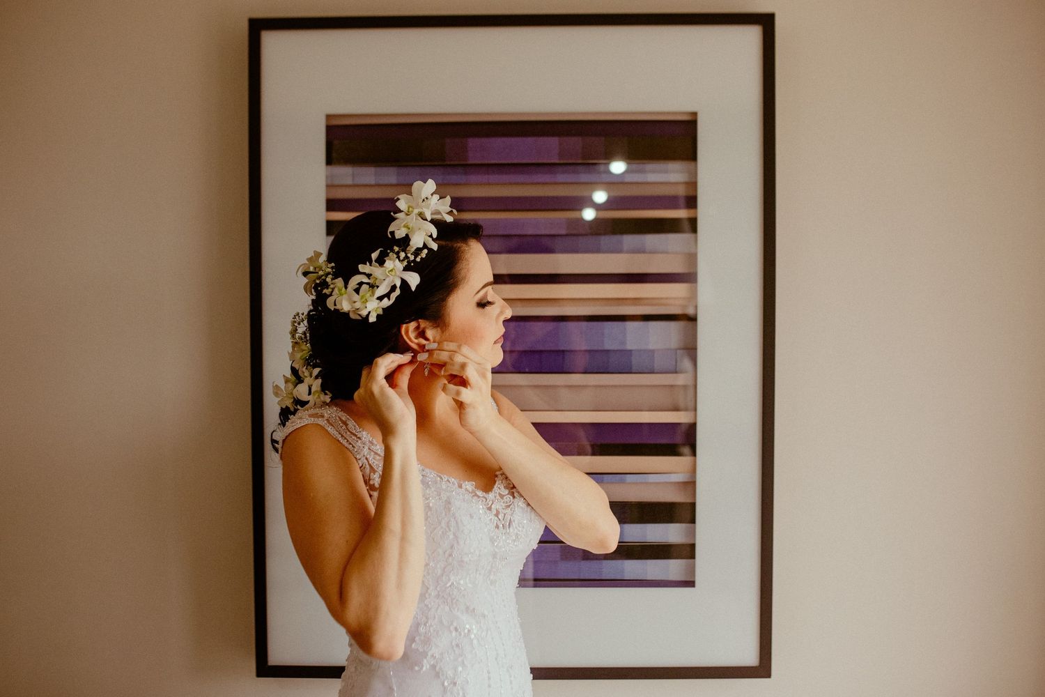 Person in wedding attire putting on earrings while standing next to a framed artwork.