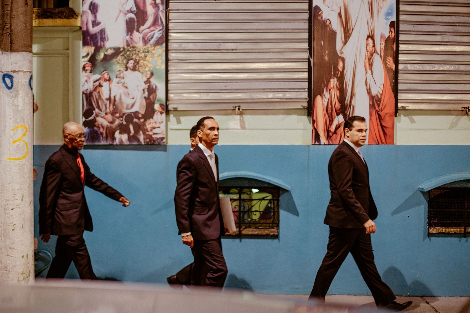 Three men in dark suits walking past a colorful mural wall with shuttered windows.
