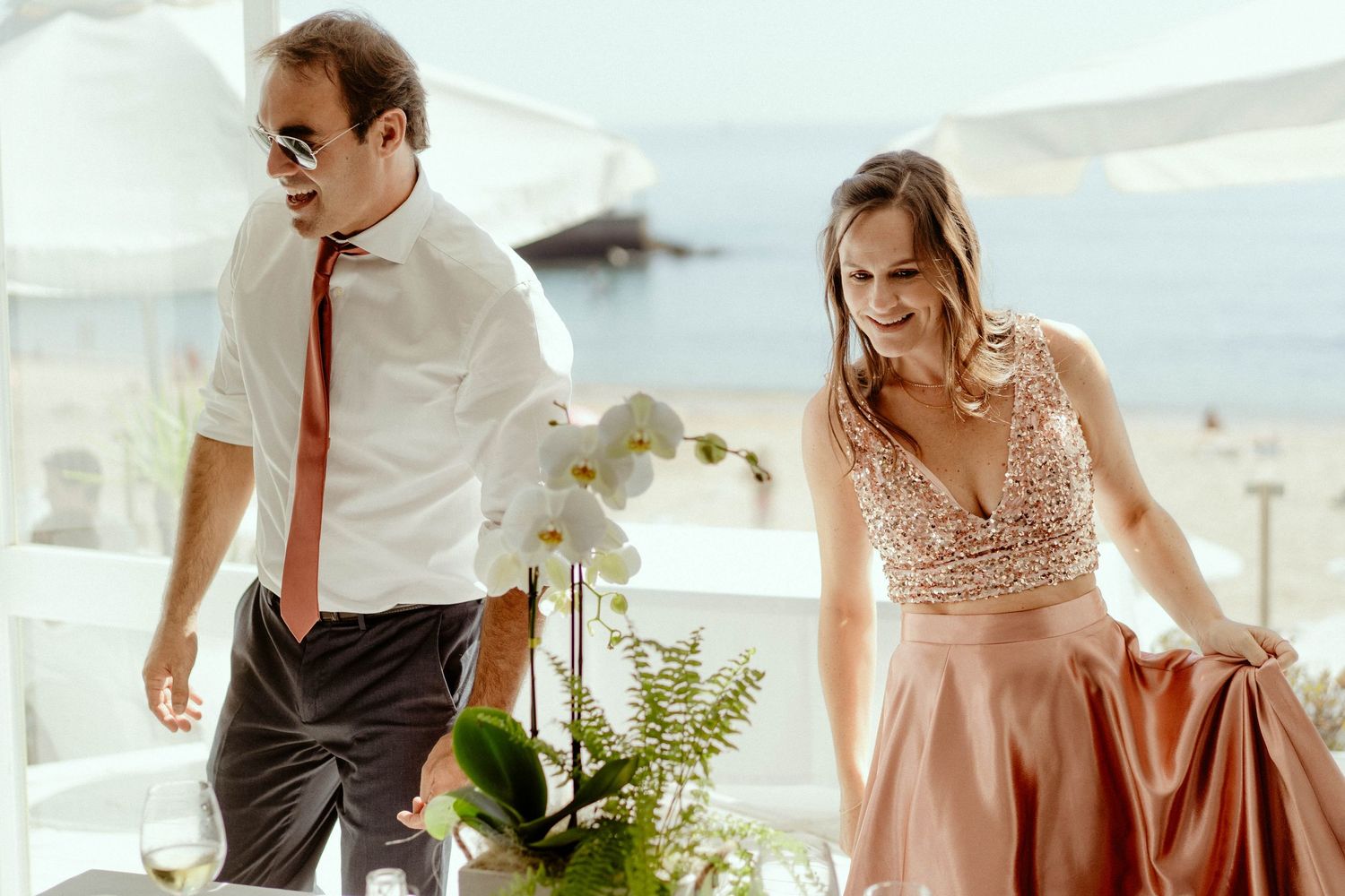 A couple in formal attire share a joyful moment at a seaside venue with white decor and tropical plants.