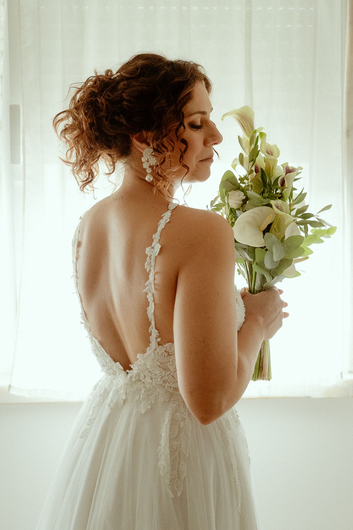 Bride in backless lace wedding dress holding white floral bouquet, photographed from behind near window.