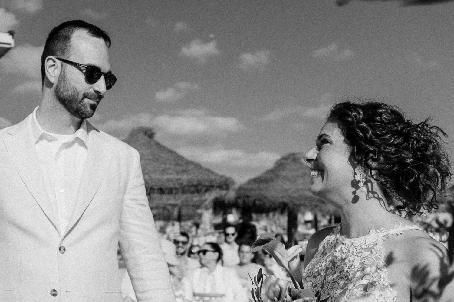 Black and white photo of a smiling bride and groom in sunglasses at an outdoor beach wedding ceremony.