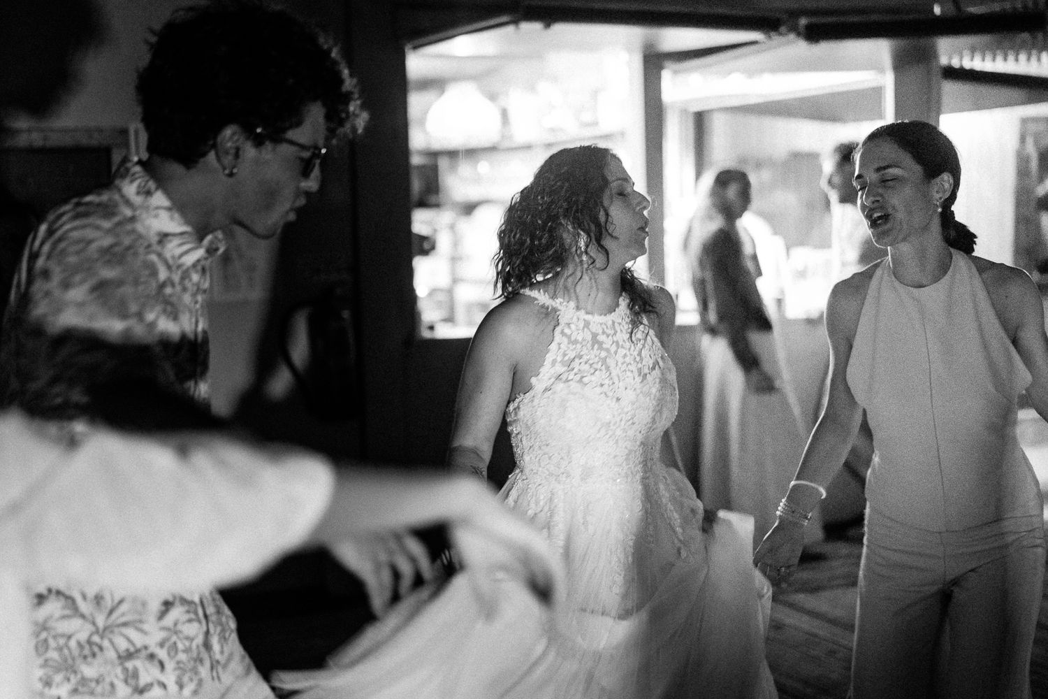 Black and white photo of a bride in a white lace dress dancing and smiling with guests at a wedding reception.