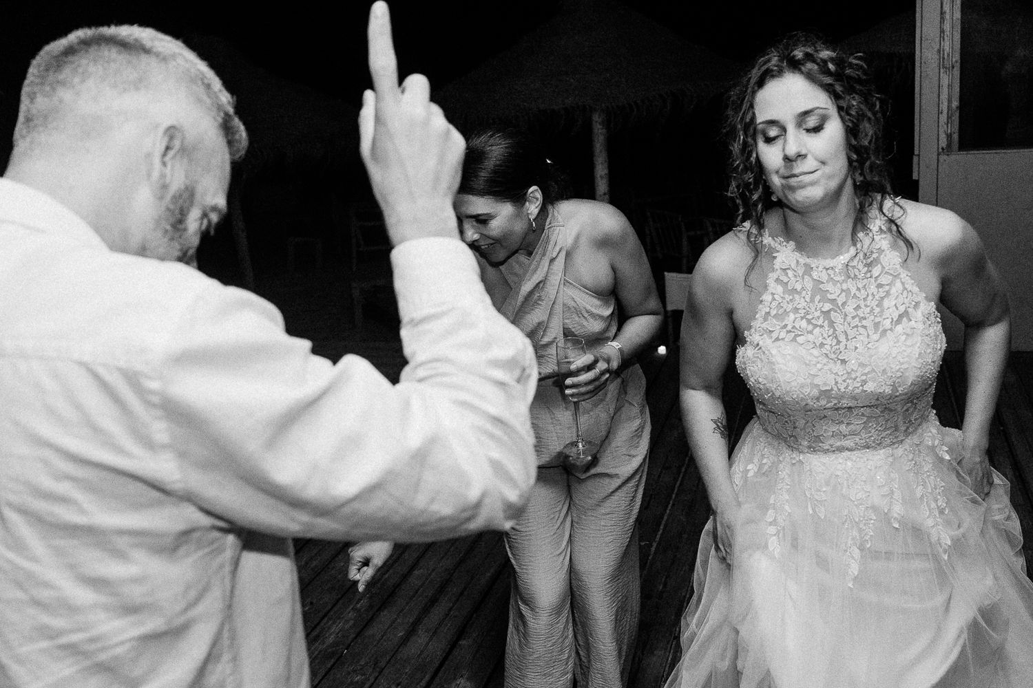 Three people dancing at a wedding reception, bride in lace gown, black and white photo.