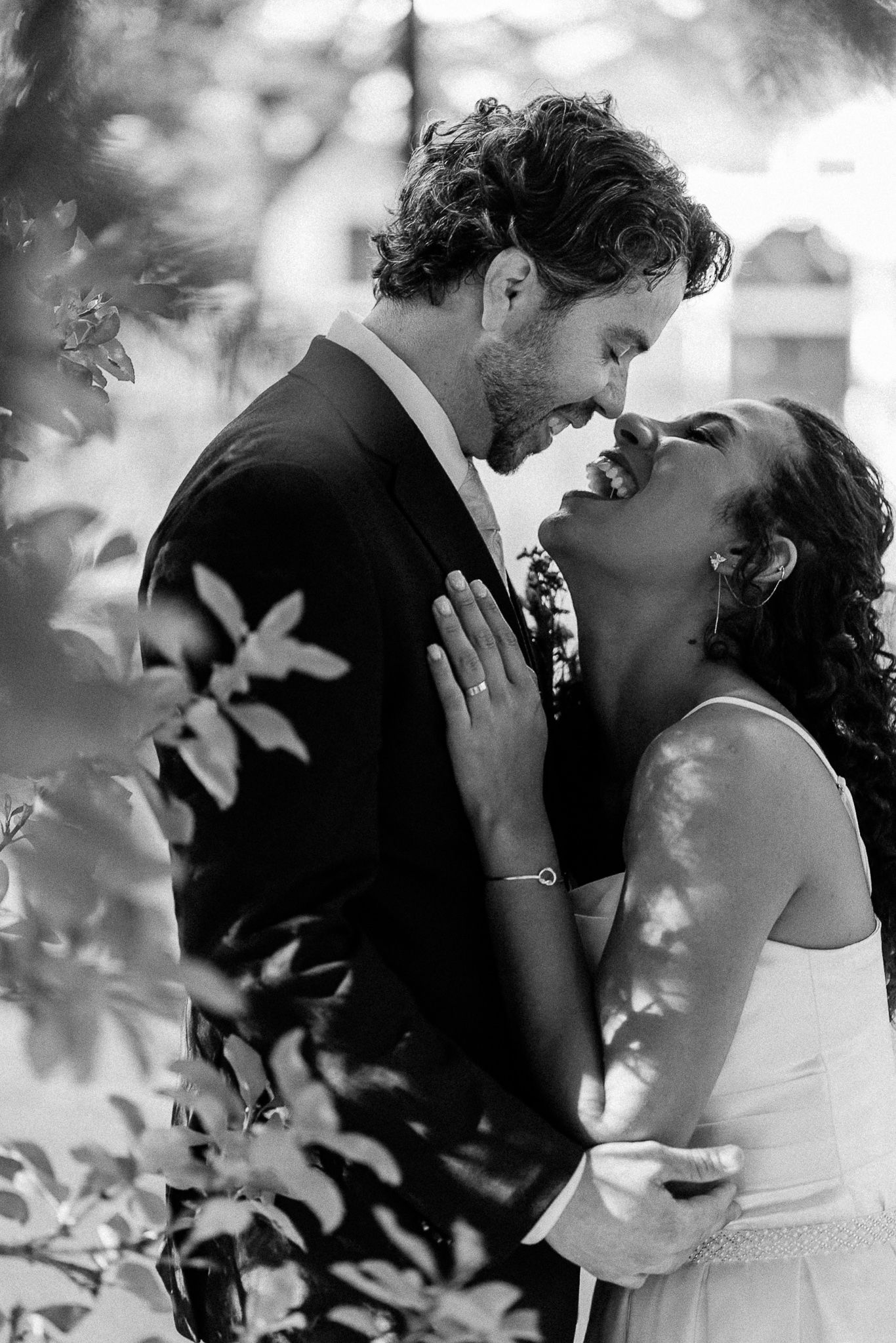 Black and white photo of a couple in wedding attire sharing a romantic moment surrounded by lush foliage.