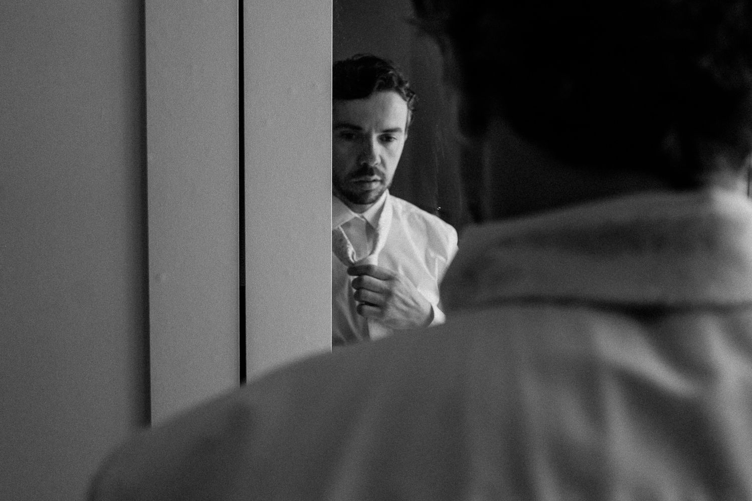 Black and white photo of a man adjusting his tie while looking in a mirror, dressed in a white shirt.