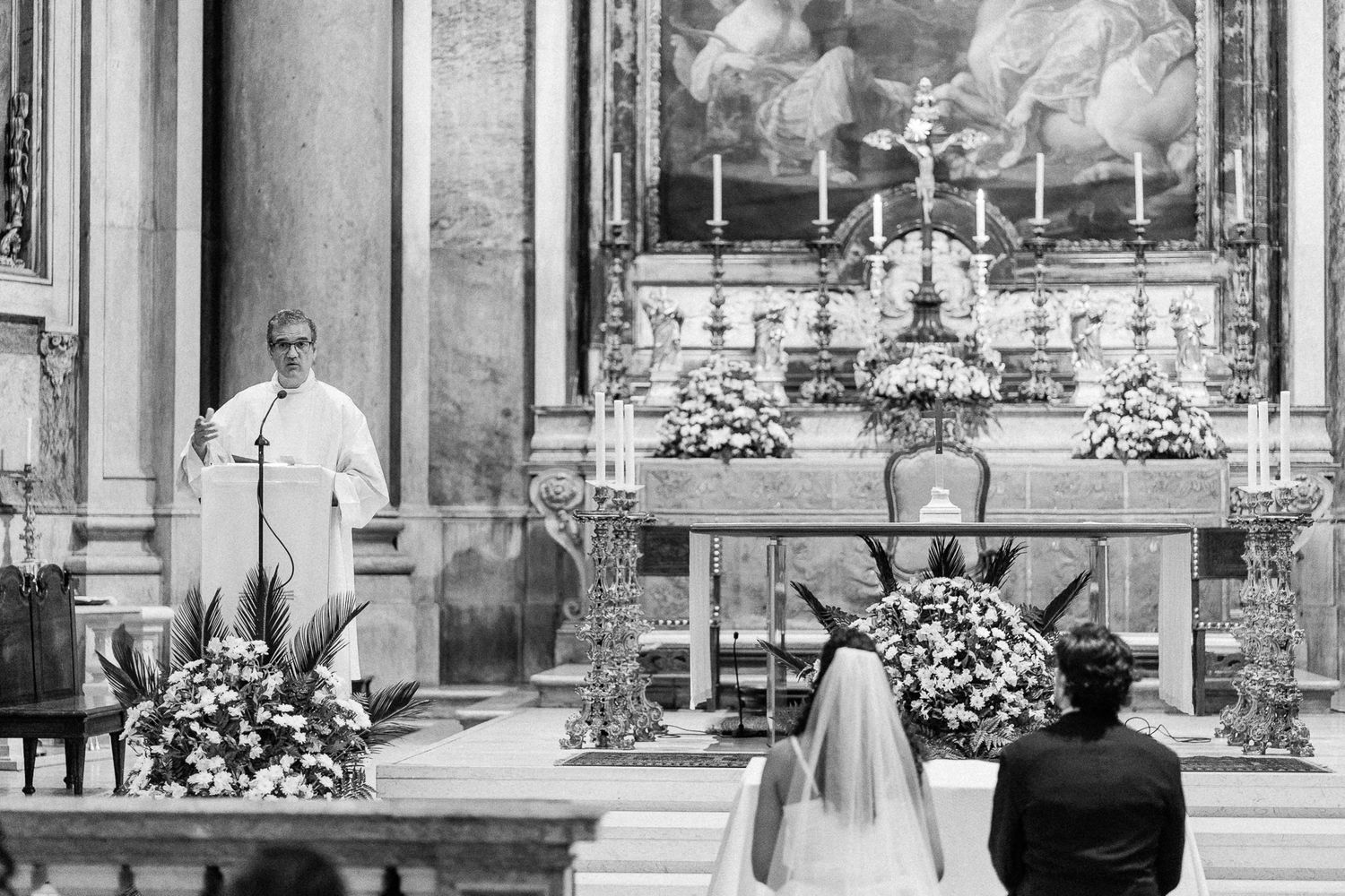 Black and white wedding ceremony photo with couple kneeling before priest at ornate church altar adorned with flowers.