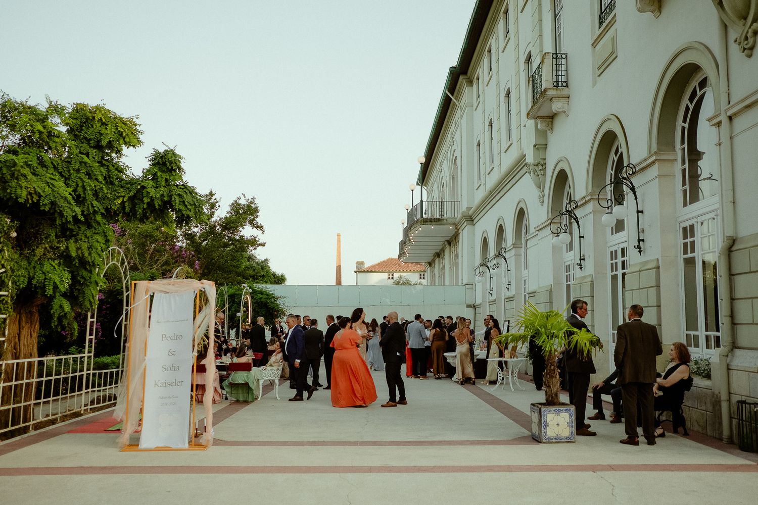Elegant outdoor event at a historic white building with guests mingling on a paved terrace at dusk.