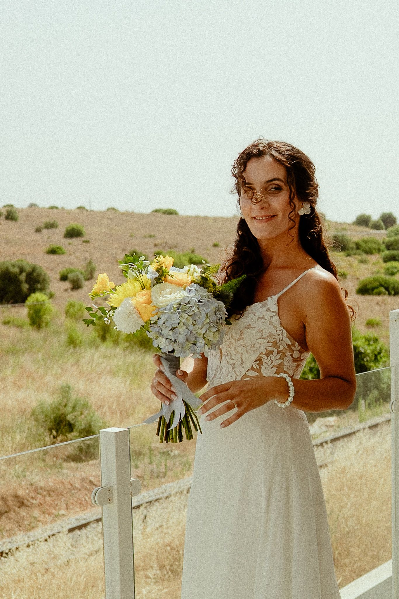 Bride in white lace dress holding yellow and blue bouquet, smiling outdoors in sunny landscape.