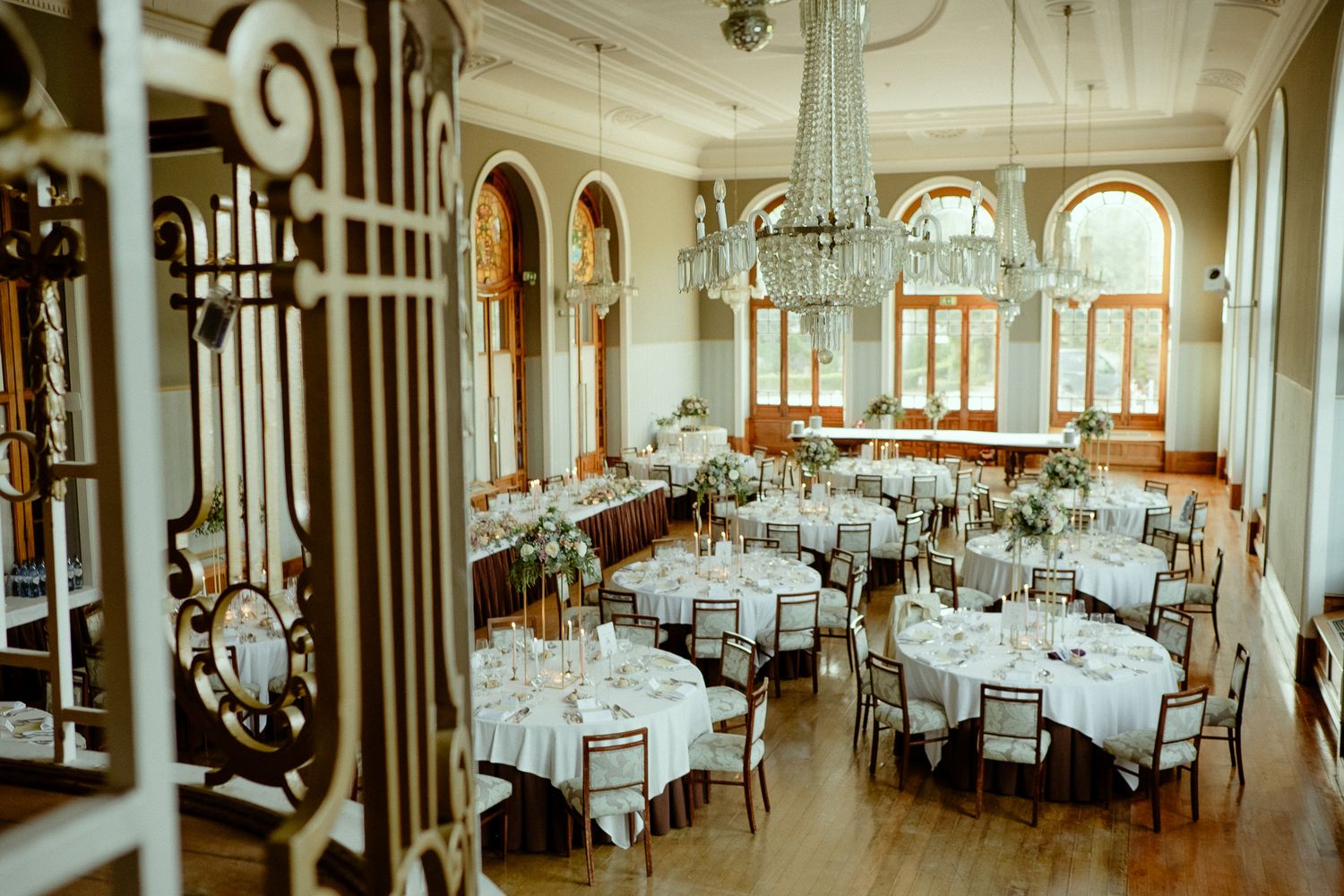 Elegant wedding reception hall with crystal chandeliers, round white-draped tables, and arched windows in a historic venue.
