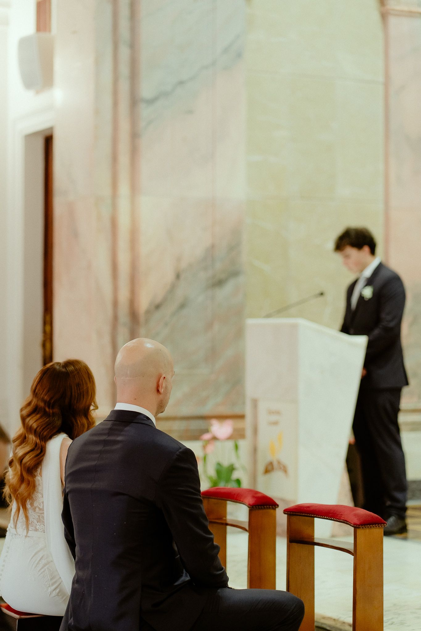 Couple seated in church pews during a wedding ceremony, with a man speaking at a podium in the background.