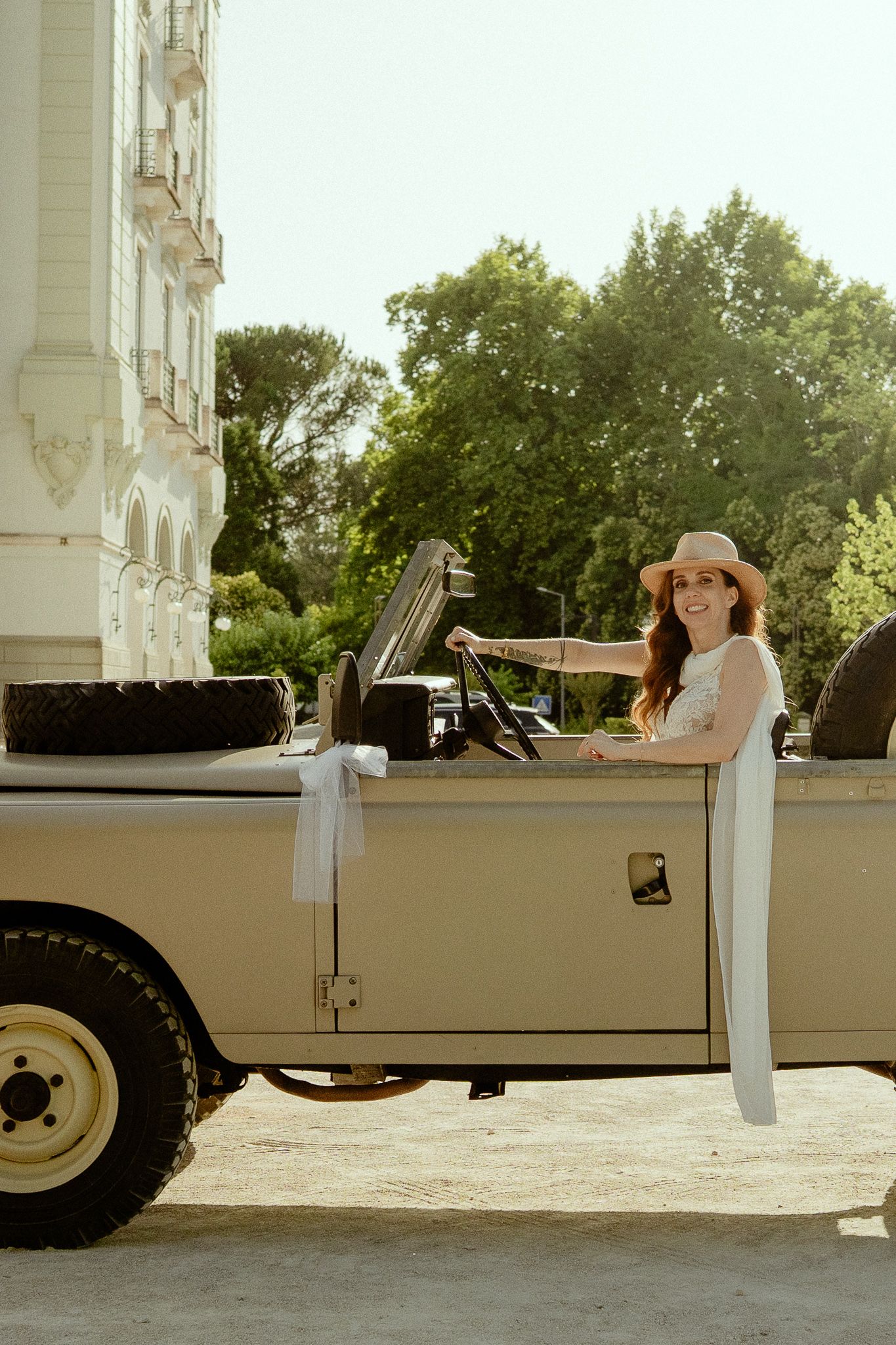 Smiling woman in white dress and hat standing beside a vintage cream convertible SUV outdoors.