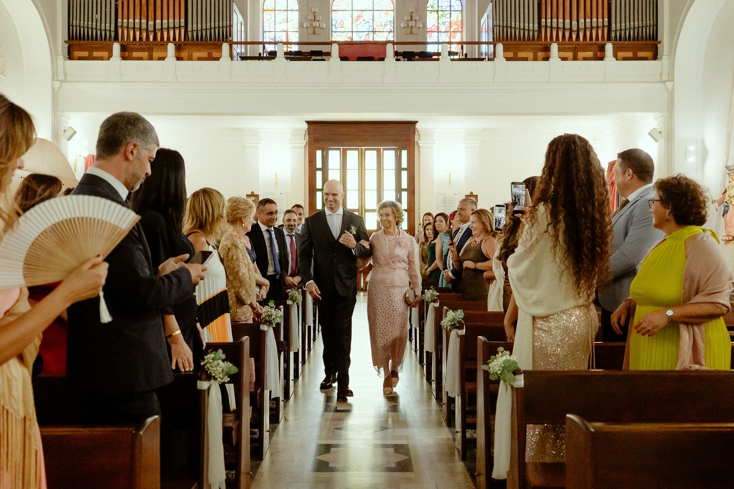 Bride walks down church aisle with escort, guests standing in pews, elegant wedding ceremony interior.