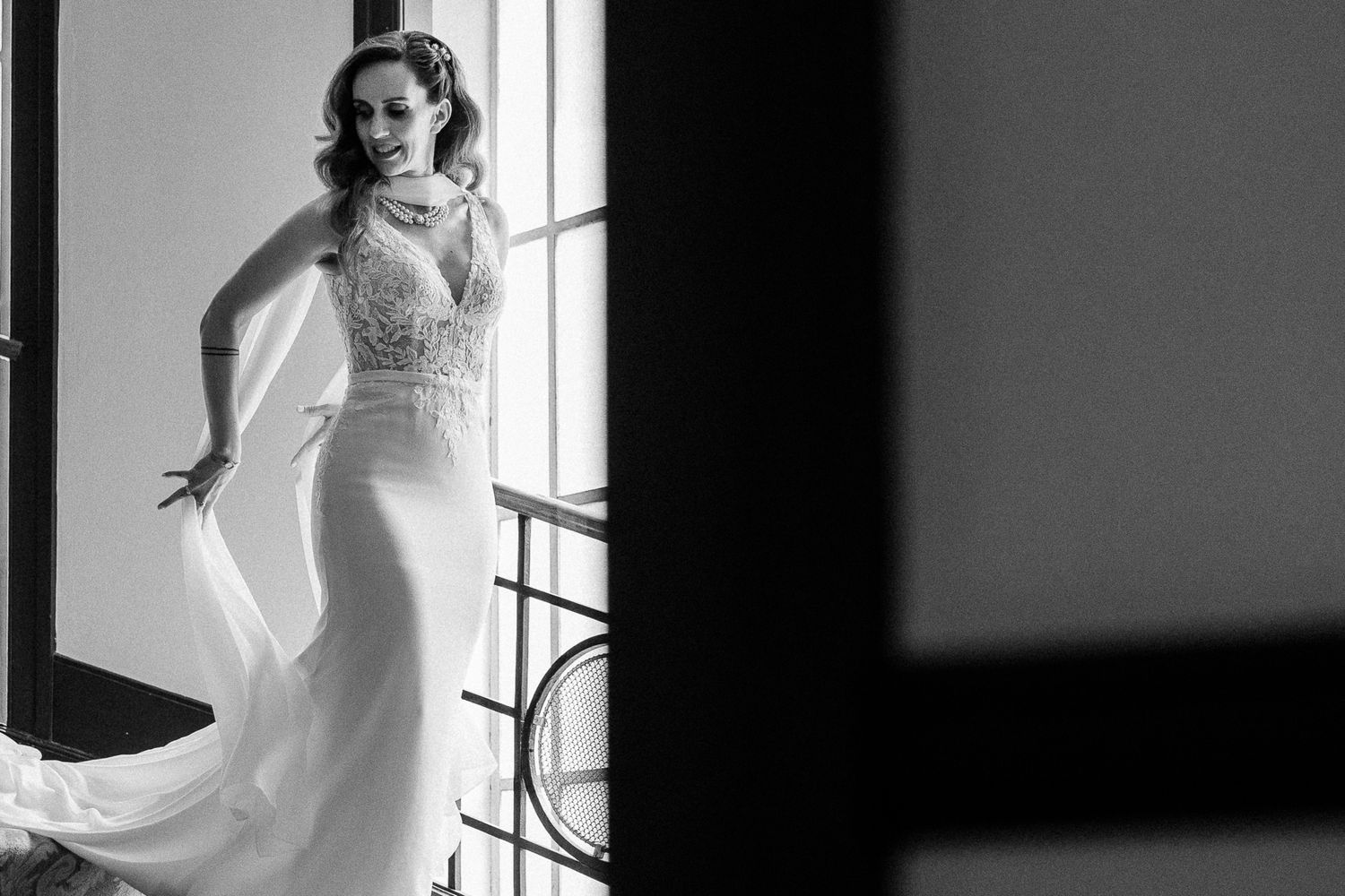 Elegant bride in a flowing white lace wedding gown posing gracefully in a doorway, black and white photography.