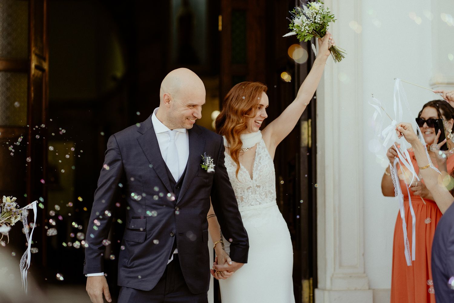Joyful newlyweds exit church as bride raises bouquet, groom in navy suit, confetti falling, guests celebrating.