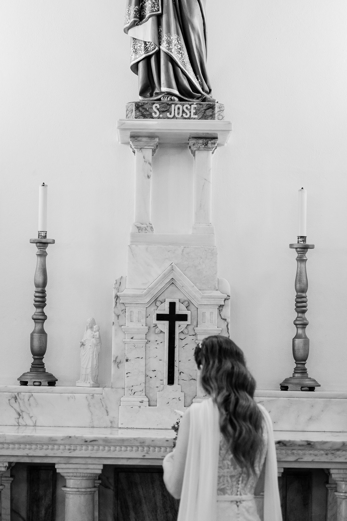 Girl in white dress praying at church altar with Saint Jose statue and candles in black and white photo.