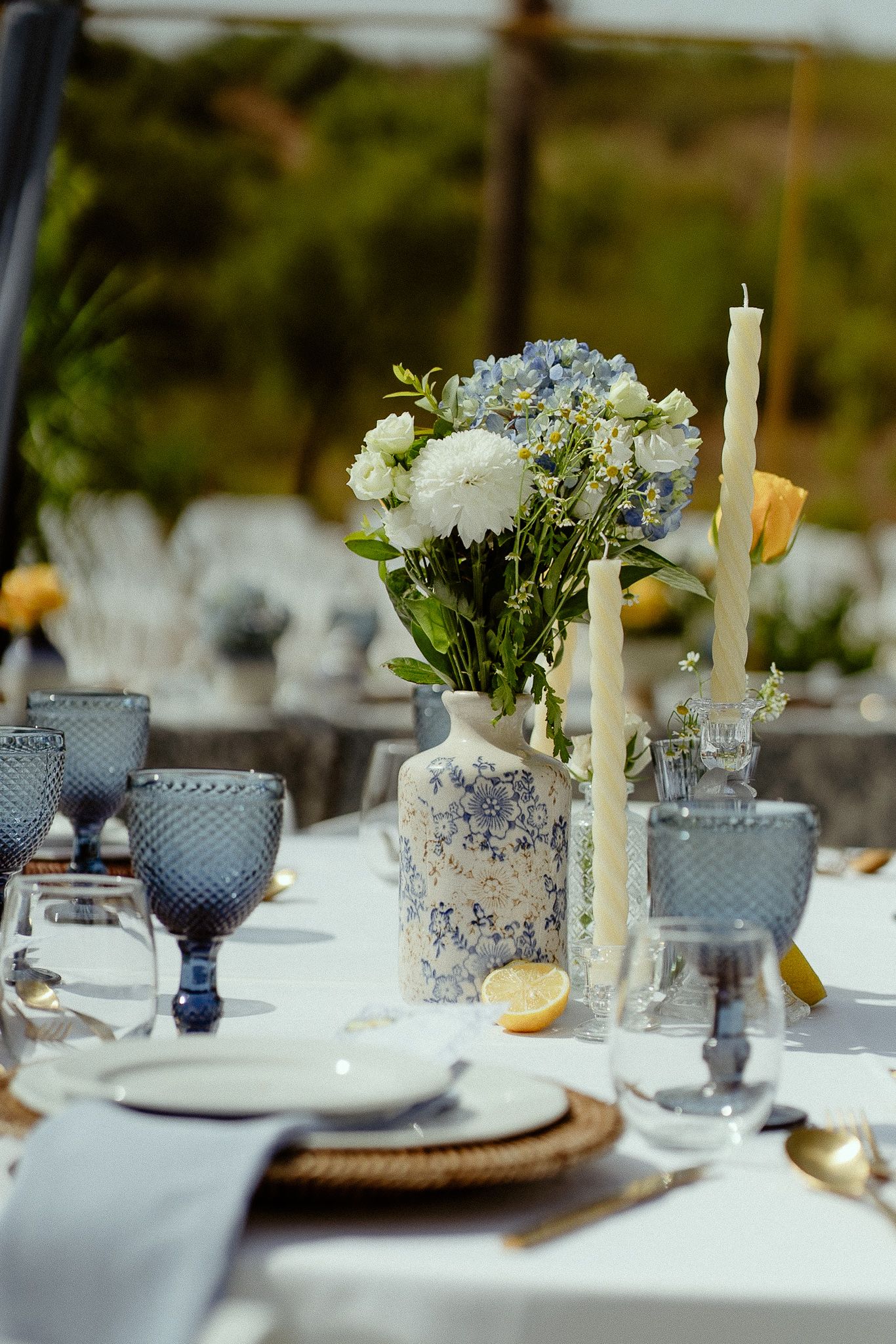 Elegant outdoor table setting with blue glassware, floral centerpiece, candles, and lemon accents on white linen.