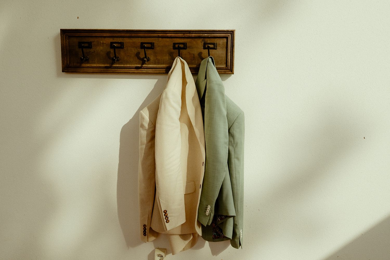 Two jackets hanging on a wooden wall-mounted coat rack against a white wall in natural light.