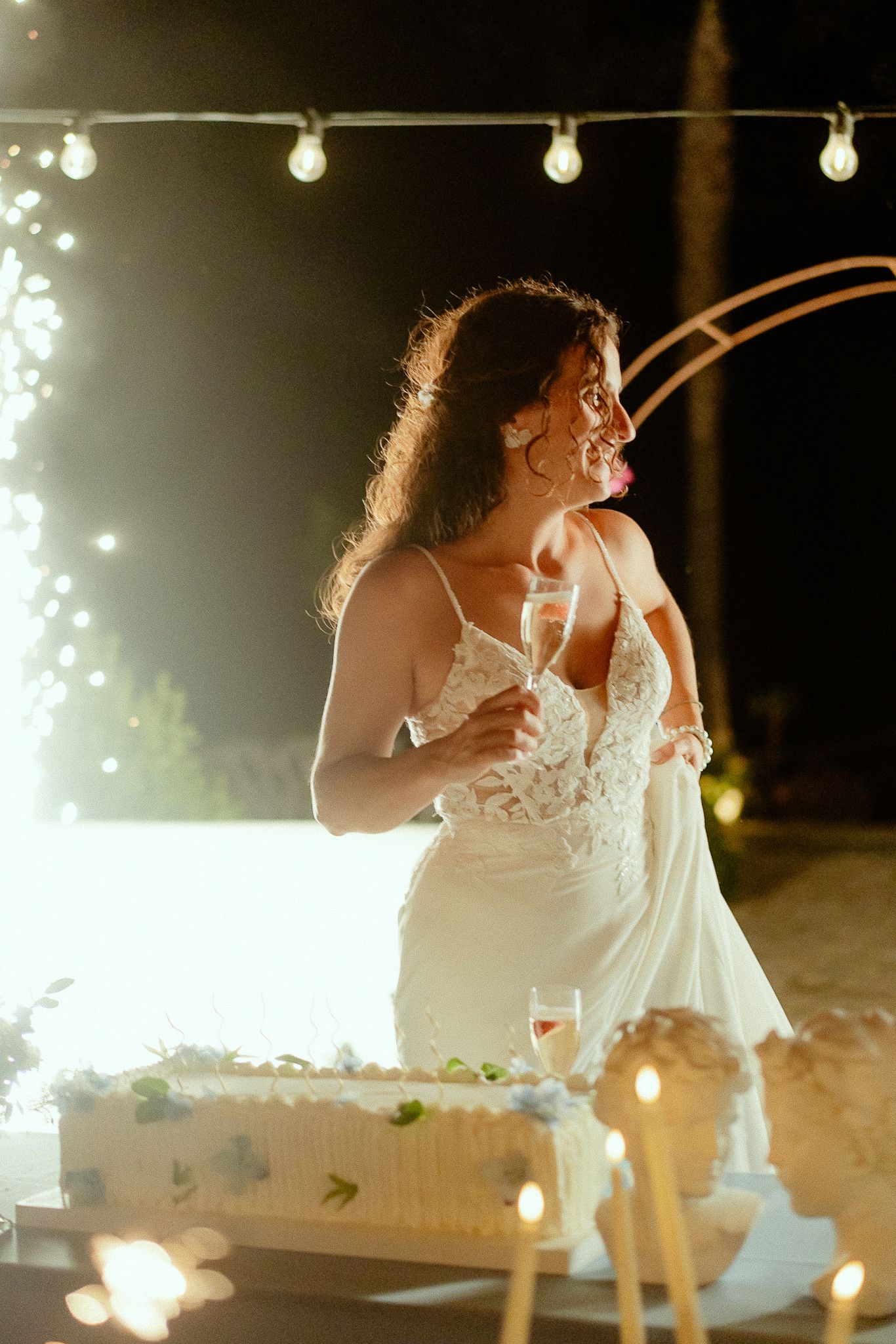 Bride in white lace gown smiling beside wedding cake with sparkling lights at night outdoor reception.