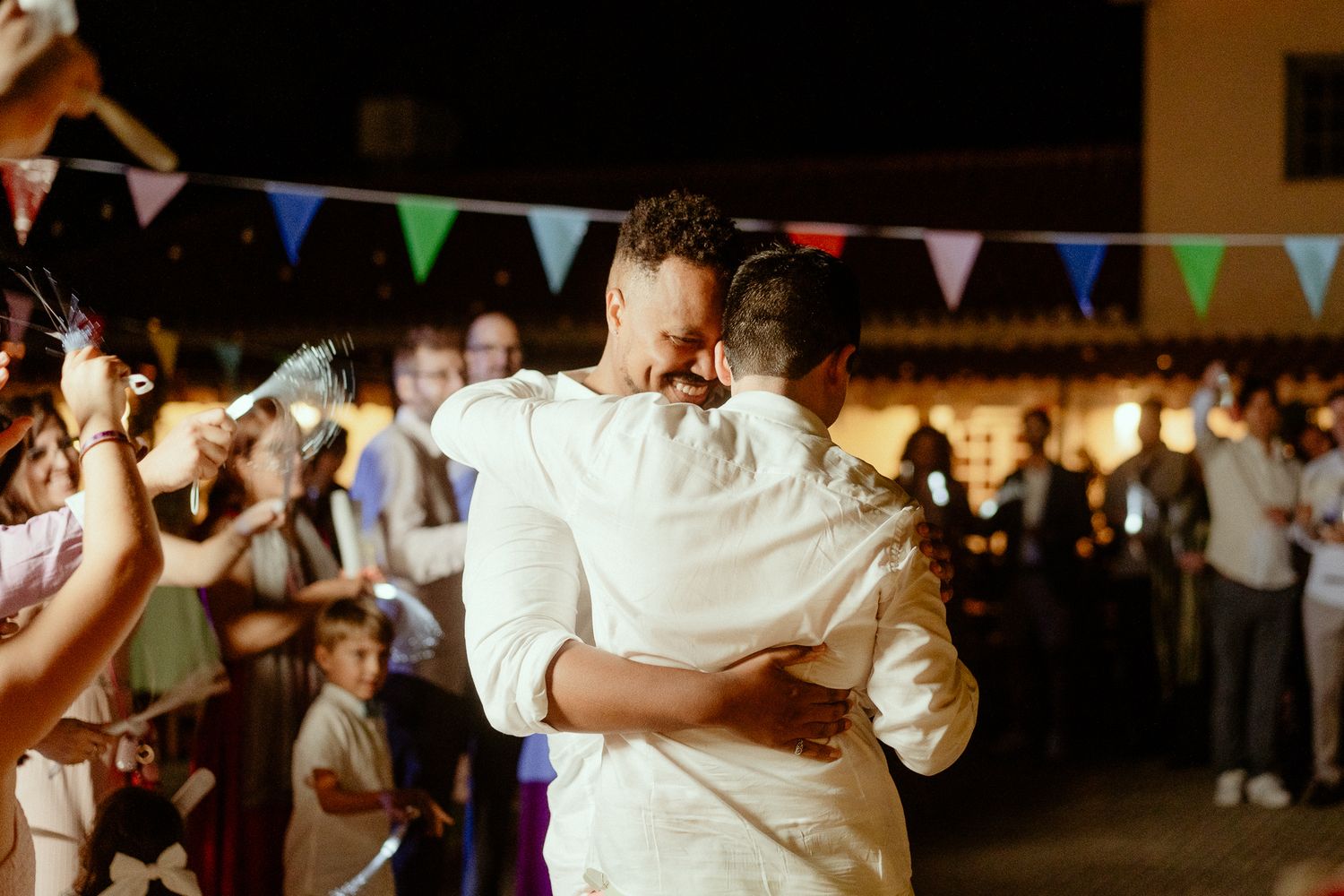 Two grooms share a first dance embrace at an outdoor wedding reception with colorful bunting lights.