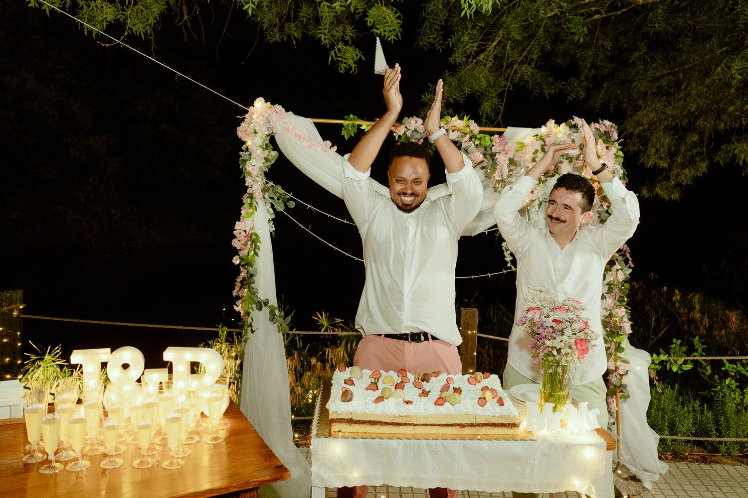 Two grooms celebrate at wedding cake cutting ceremony with floral arch and illuminated TR initials at night.