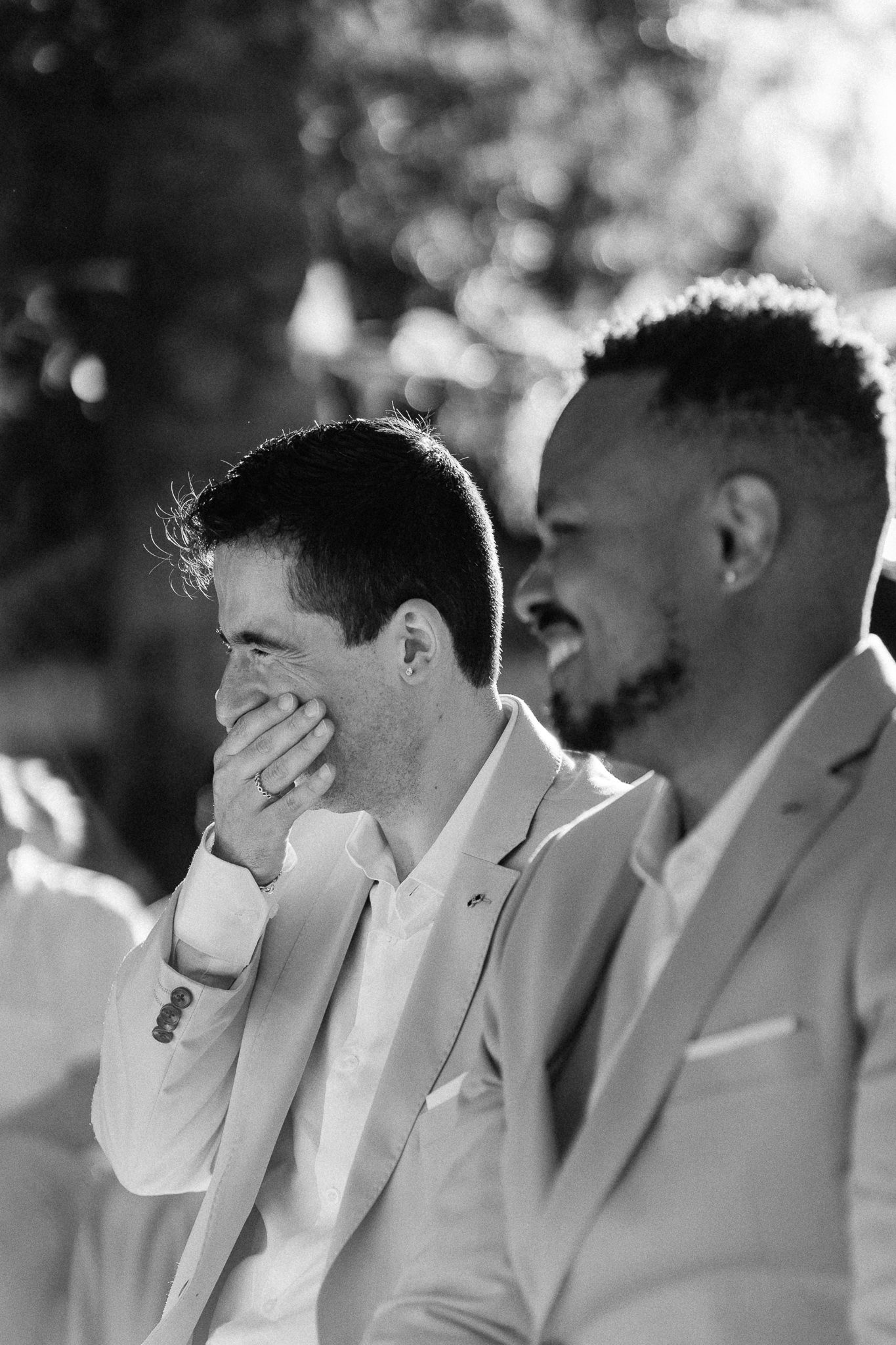 Black and white photo of two men in suits laughing joyfully at a wedding ceremony outdoors.