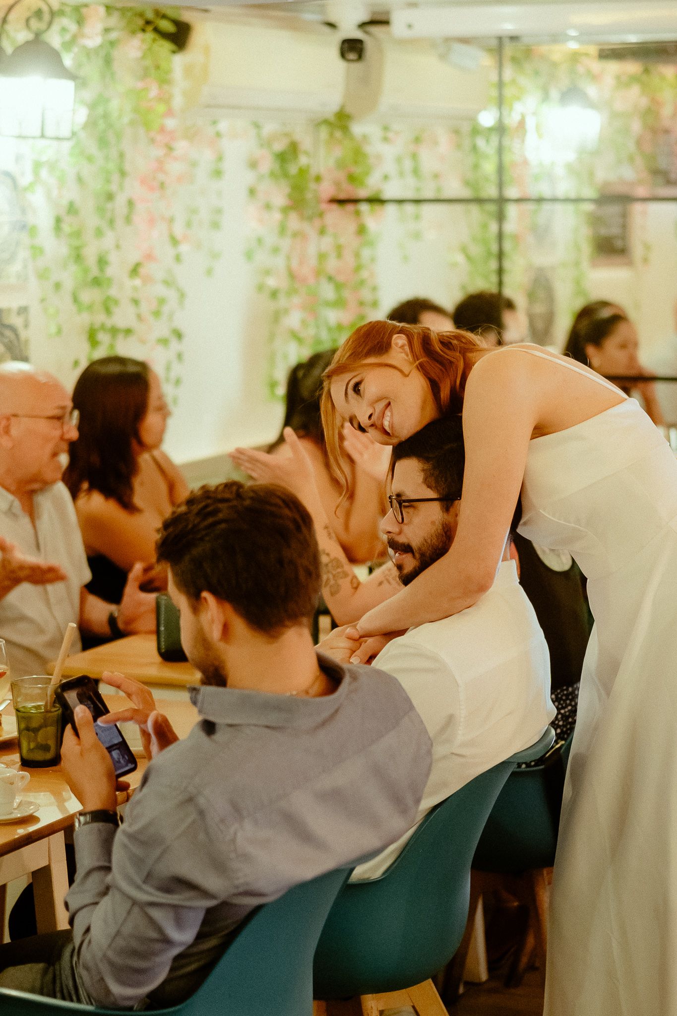 Bride in white dress hugging a seated guest at a wedding reception in a floral-decorated venue.