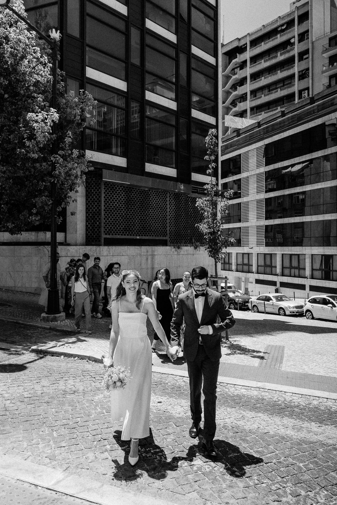 Black and white photo of a bride and groom walking hand in hand on cobblestone street with modern buildings backdrop.