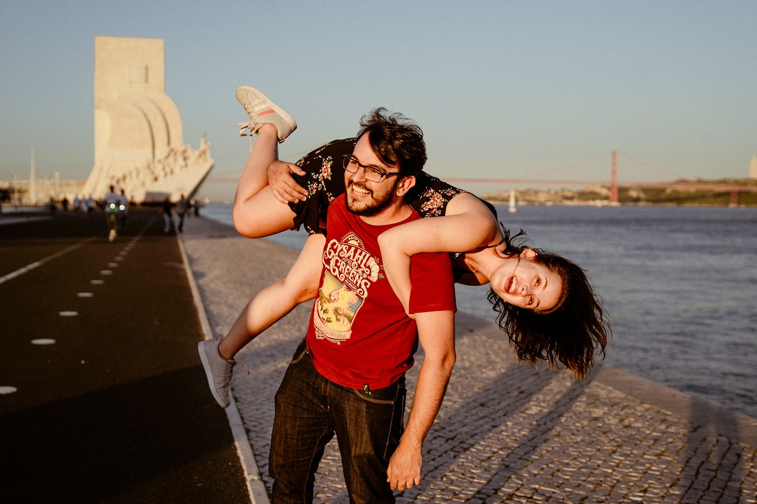 Two people playfully interacting on a waterfront promenade at sunset with a landmark monument visible in the background.