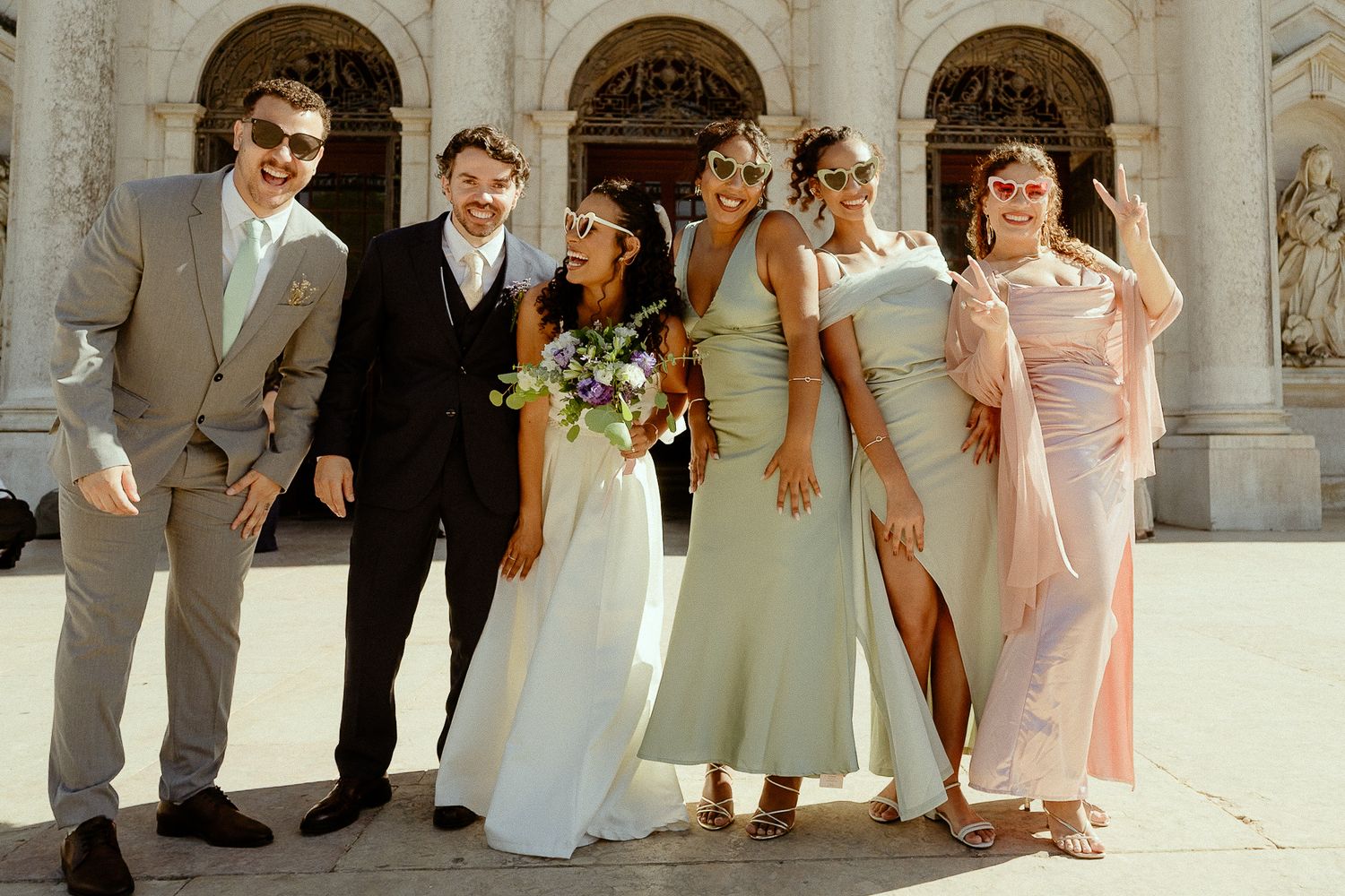 Wedding party posing joyfully outside historic building, bride in white gown with bridesmaids and groomsmen in elegant attire.
