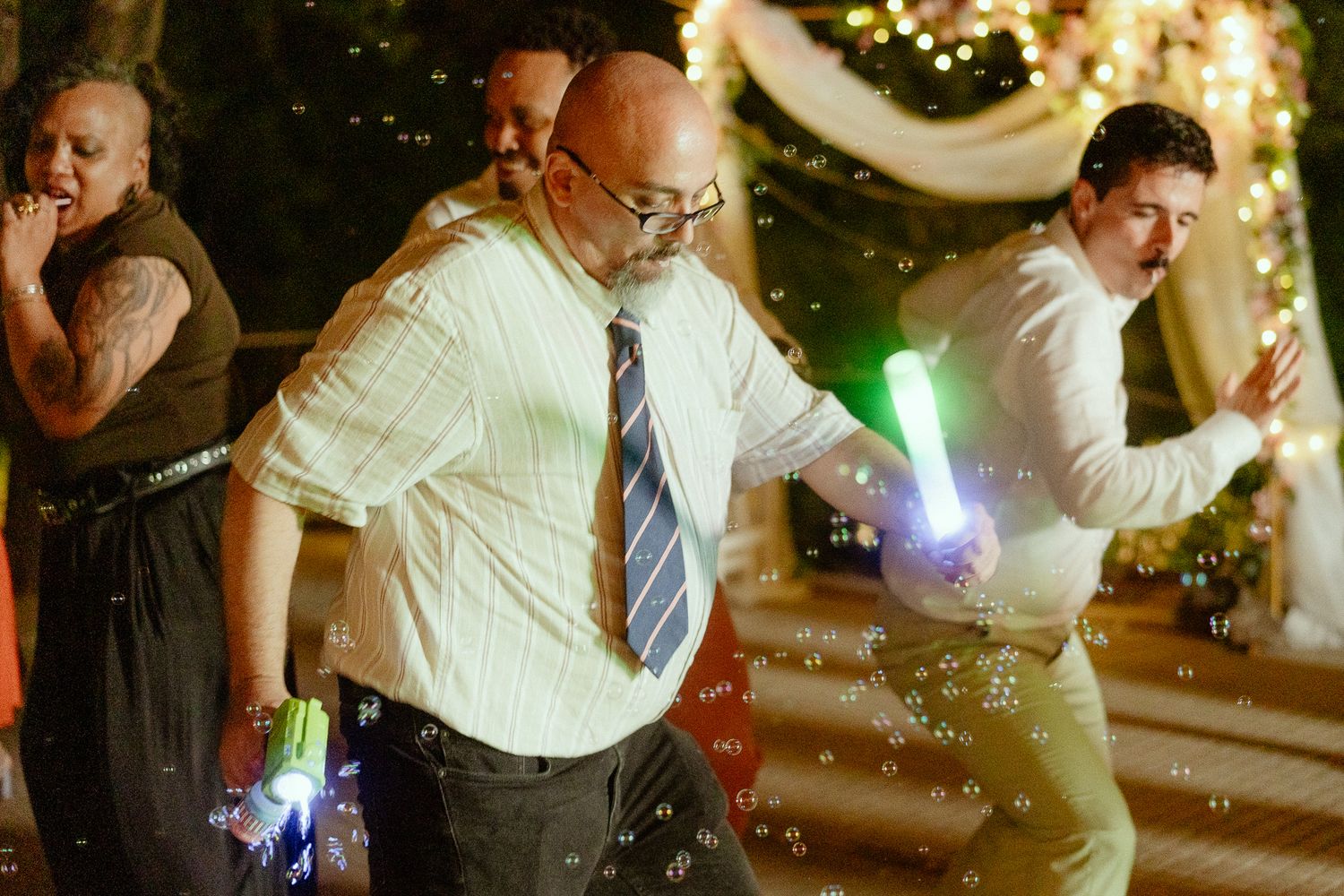 Adults dancing joyfully at a wedding reception, holding glowing light sticks amid floating bubbles at night.