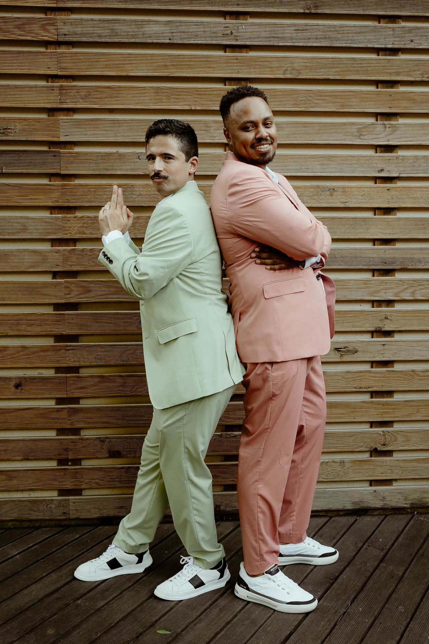 Two stylish men in pastel suits posing back-to-back against a wooden wall, smiling confidently.