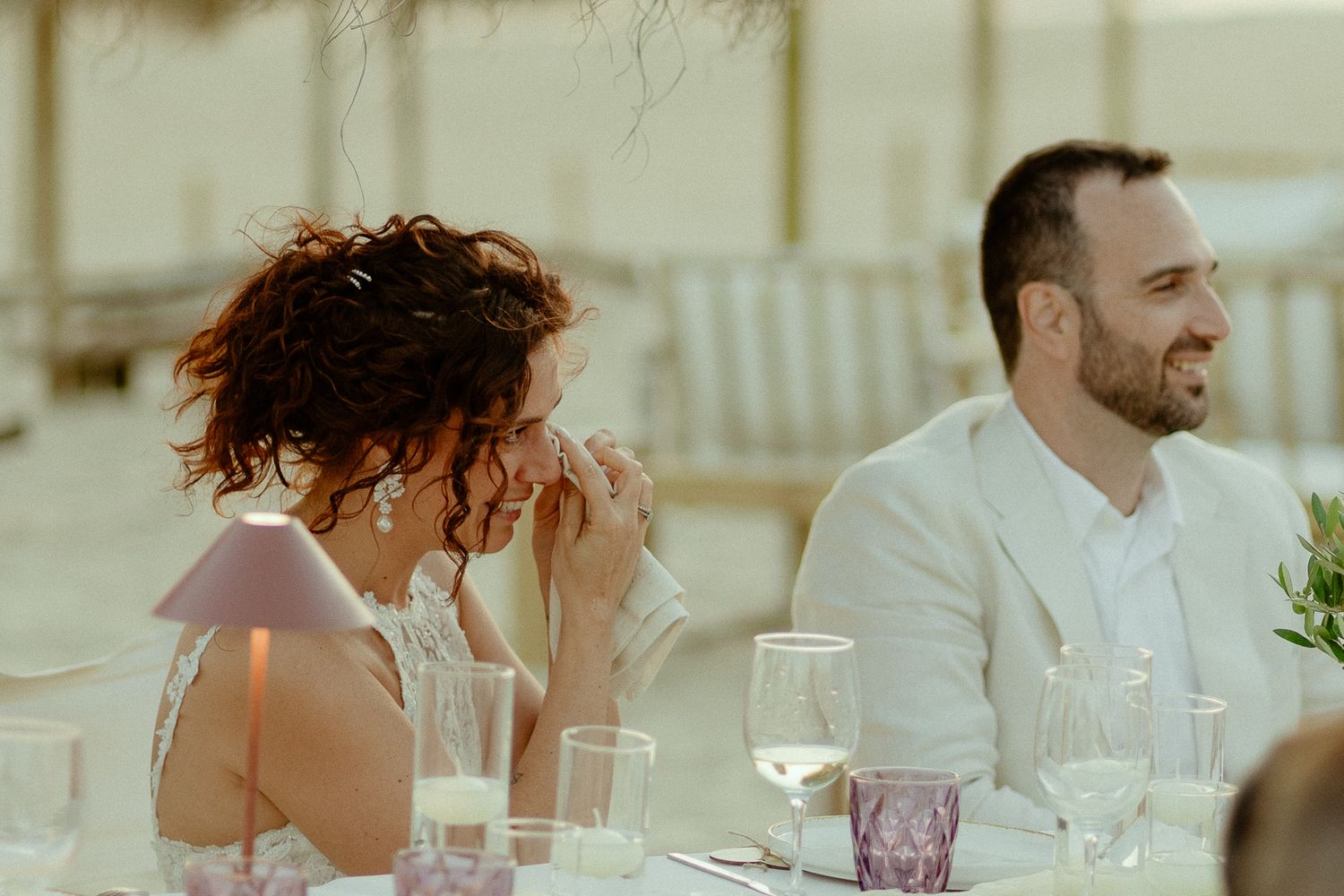 Bride and groom laughing at wedding reception table with elegant glassware at a beach venue.