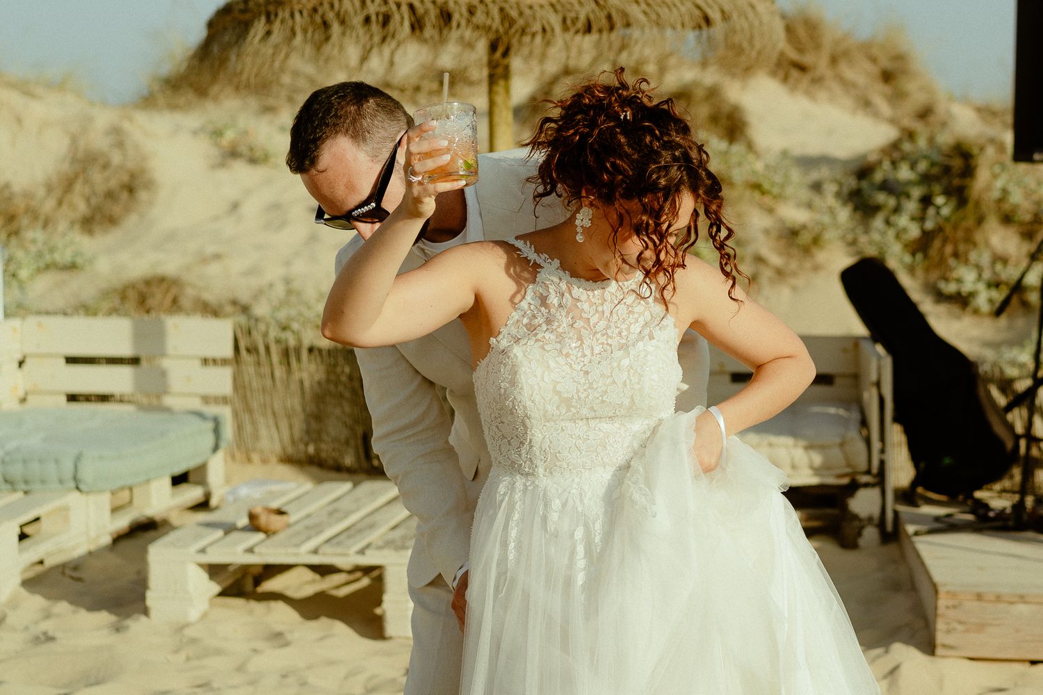 A bride in a white lace gown and groom in gray suit share a drink on a sandy beach wedding celebration.