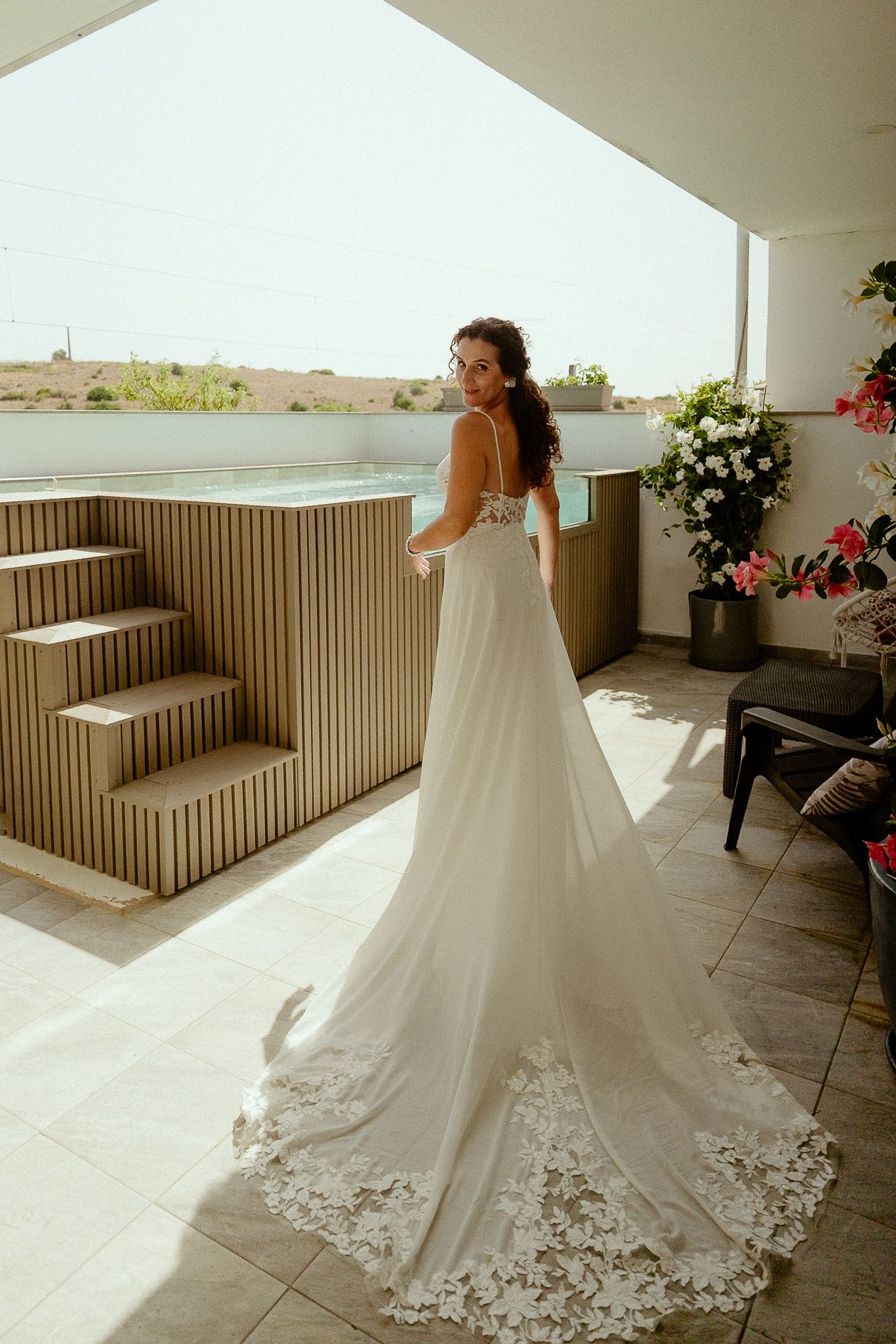 Bride in elegant white lace wedding gown with long train stands on sunny terrace, looking back over shoulder.