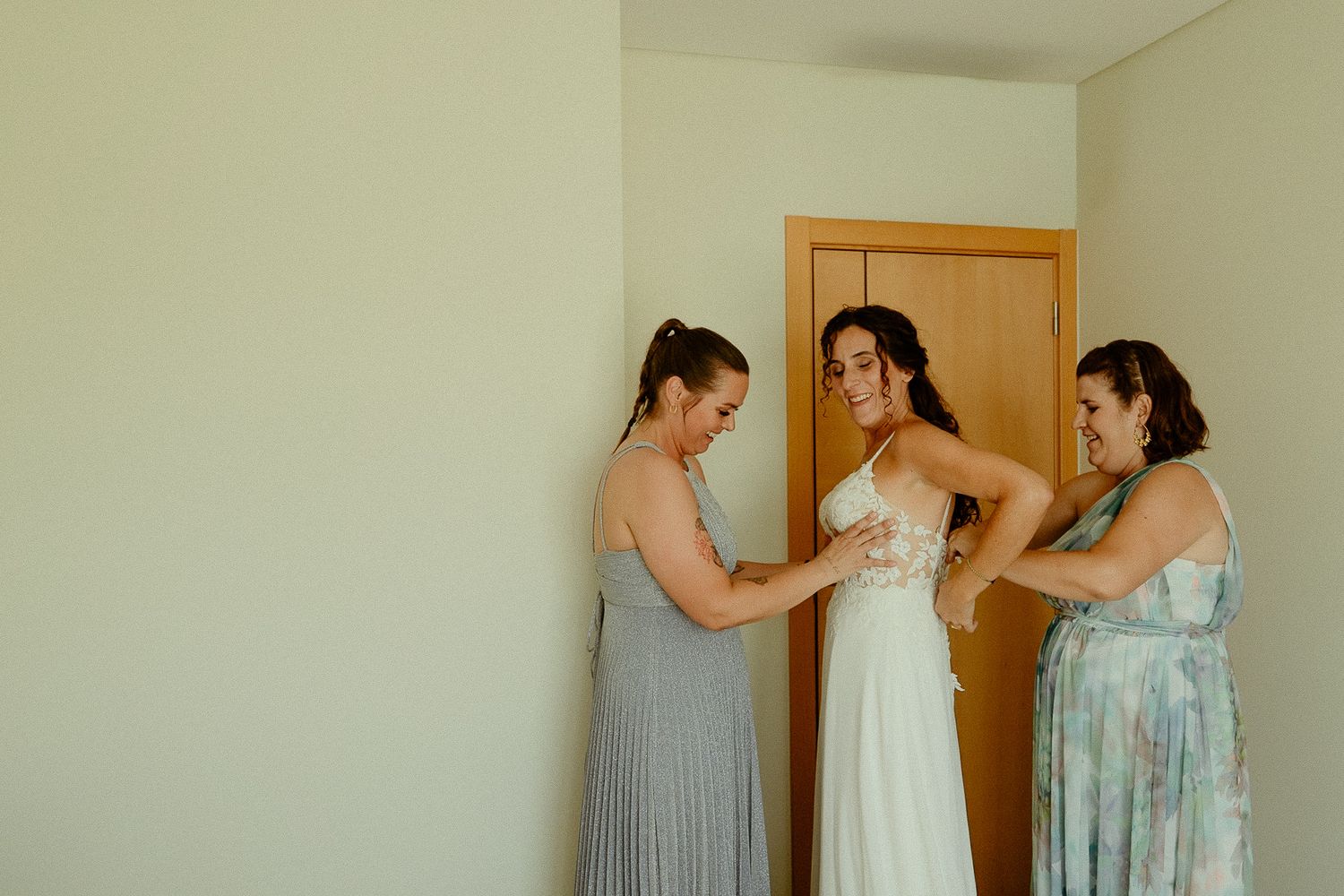 Bride being helped into white wedding dress by two bridesmaids in floral and gray gowns in a bright room.