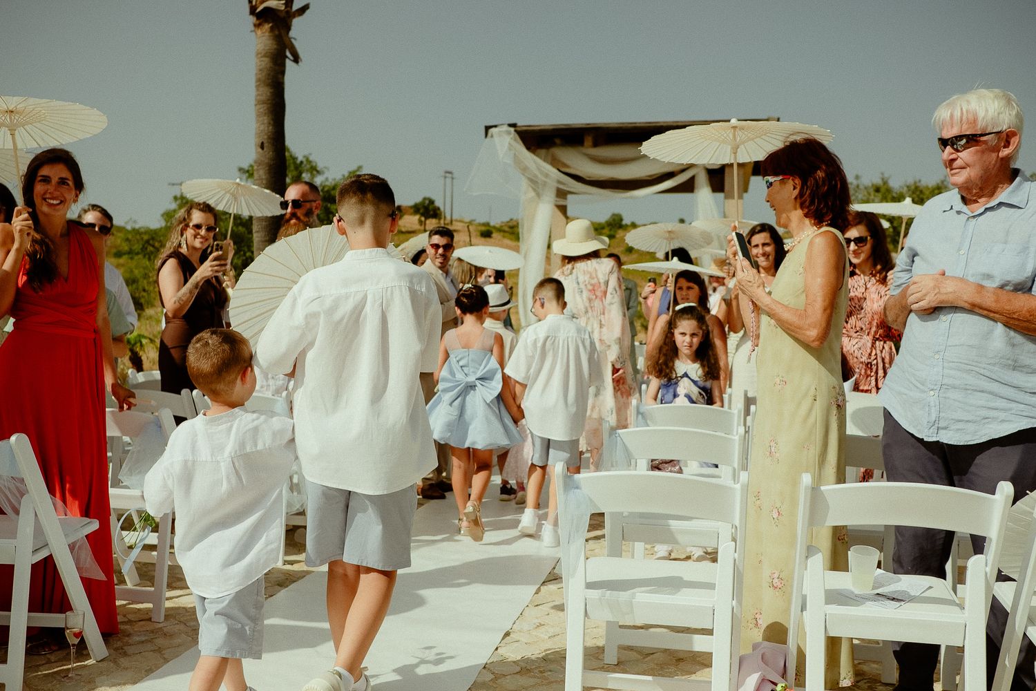 Guests celebrate as couple walks down aisle at outdoor beach wedding ceremony with white chairs and floral arch.