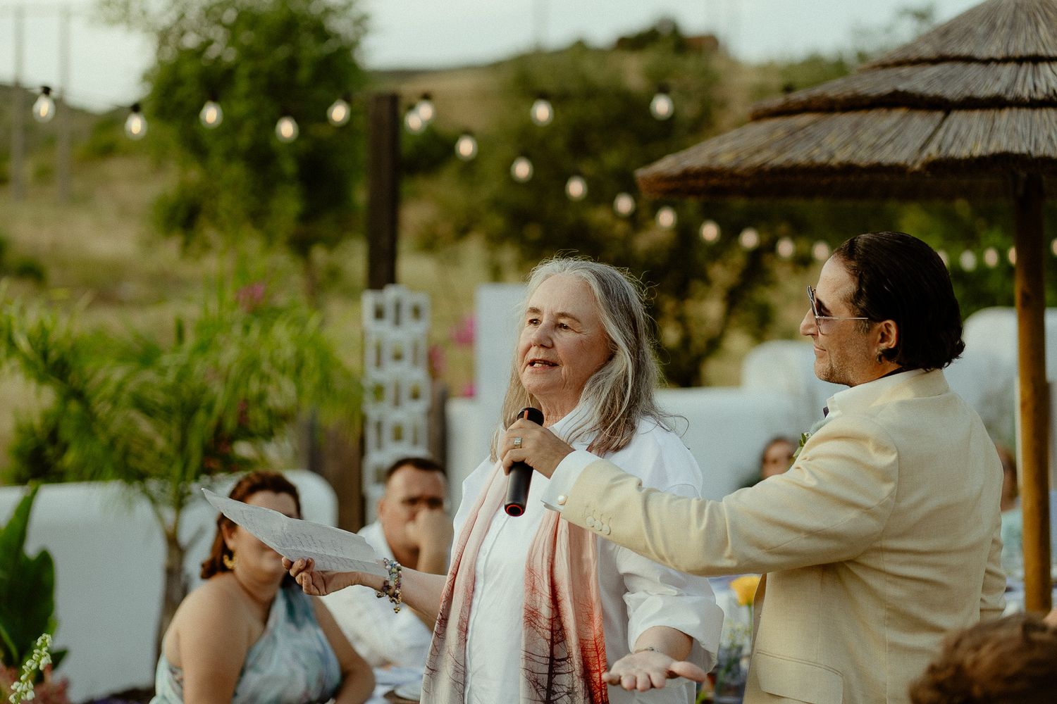 Elderly woman speaking into microphone at outdoor wedding ceremony with string lights and guests in background.