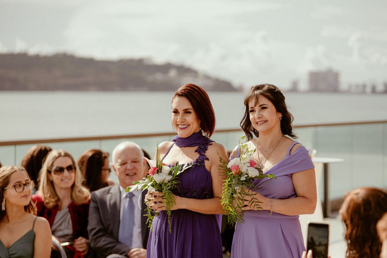 Bridesmaids in purple dresses hold bouquets during an outdoor waterfront wedding ceremony.