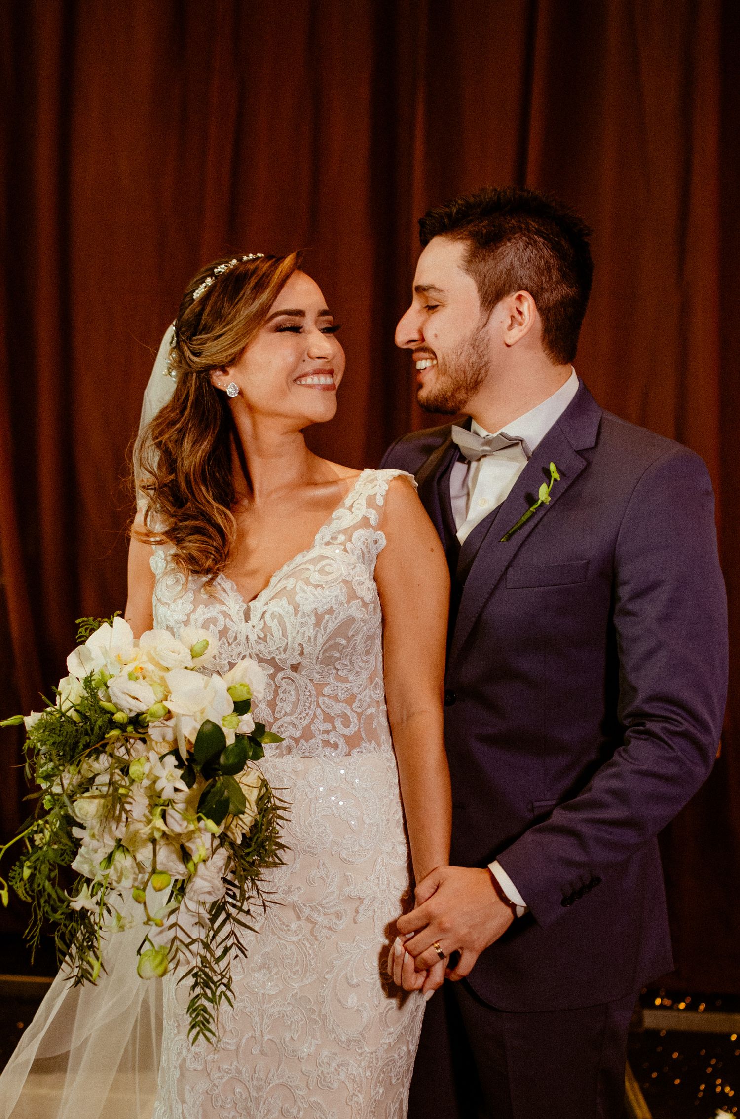 A couple at their wedding ceremony shares a tender moment in front of a brown curtain backdrop.